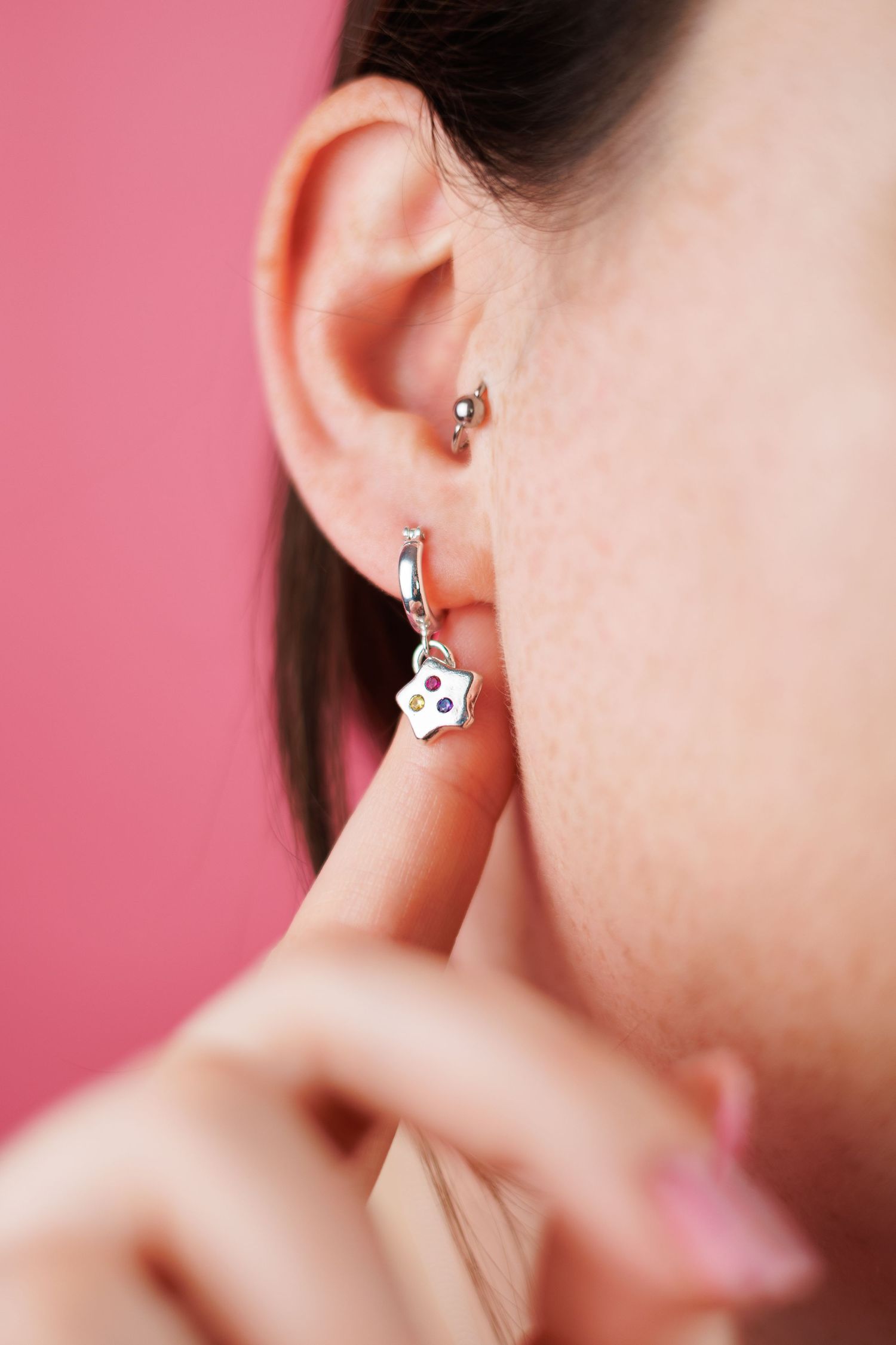 Close-up of womans ear with silver star charm hoop earring on pink background.