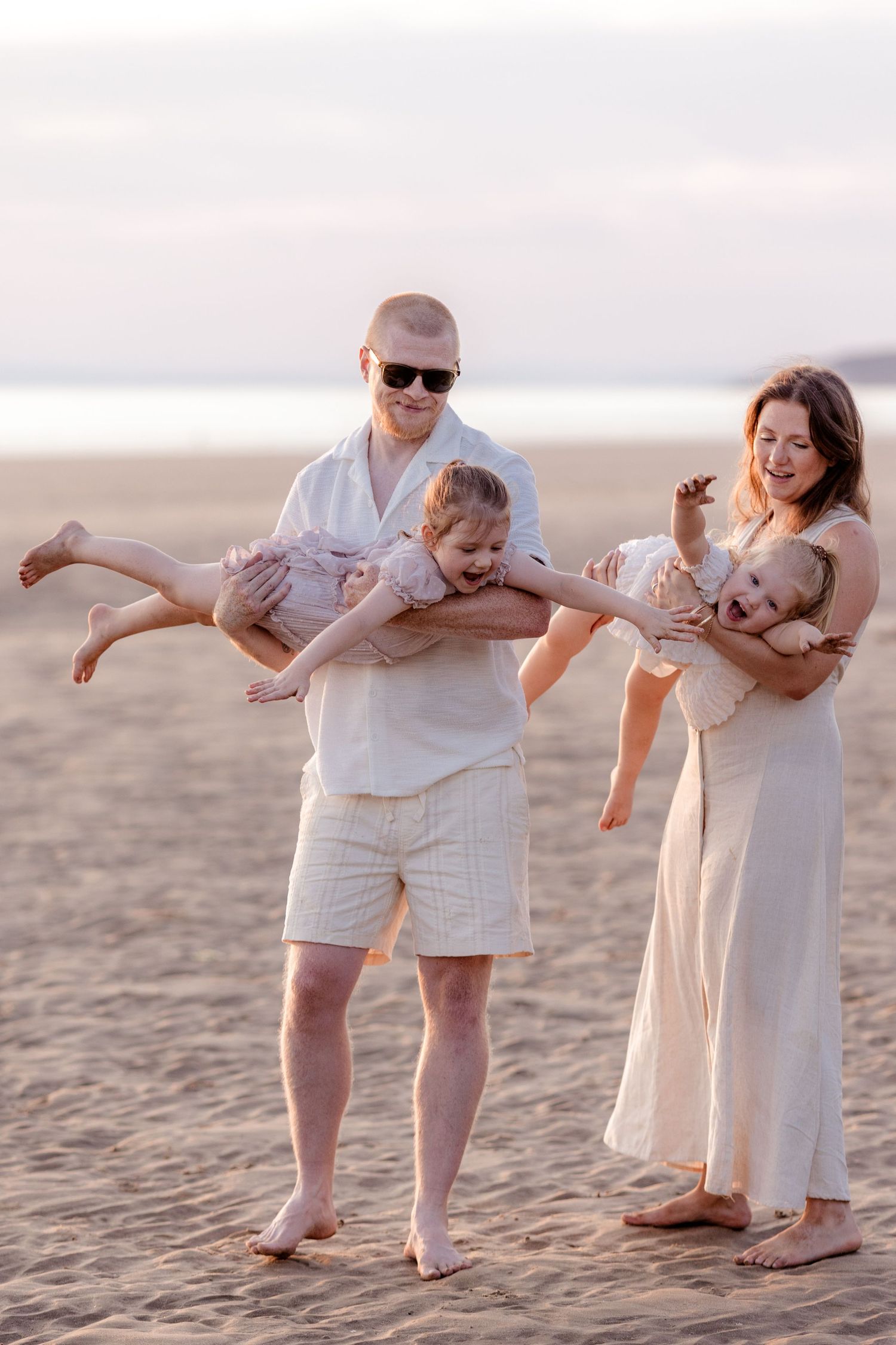 Happy family with two young children playing on a sandy beach at sunset, all dressed in white.