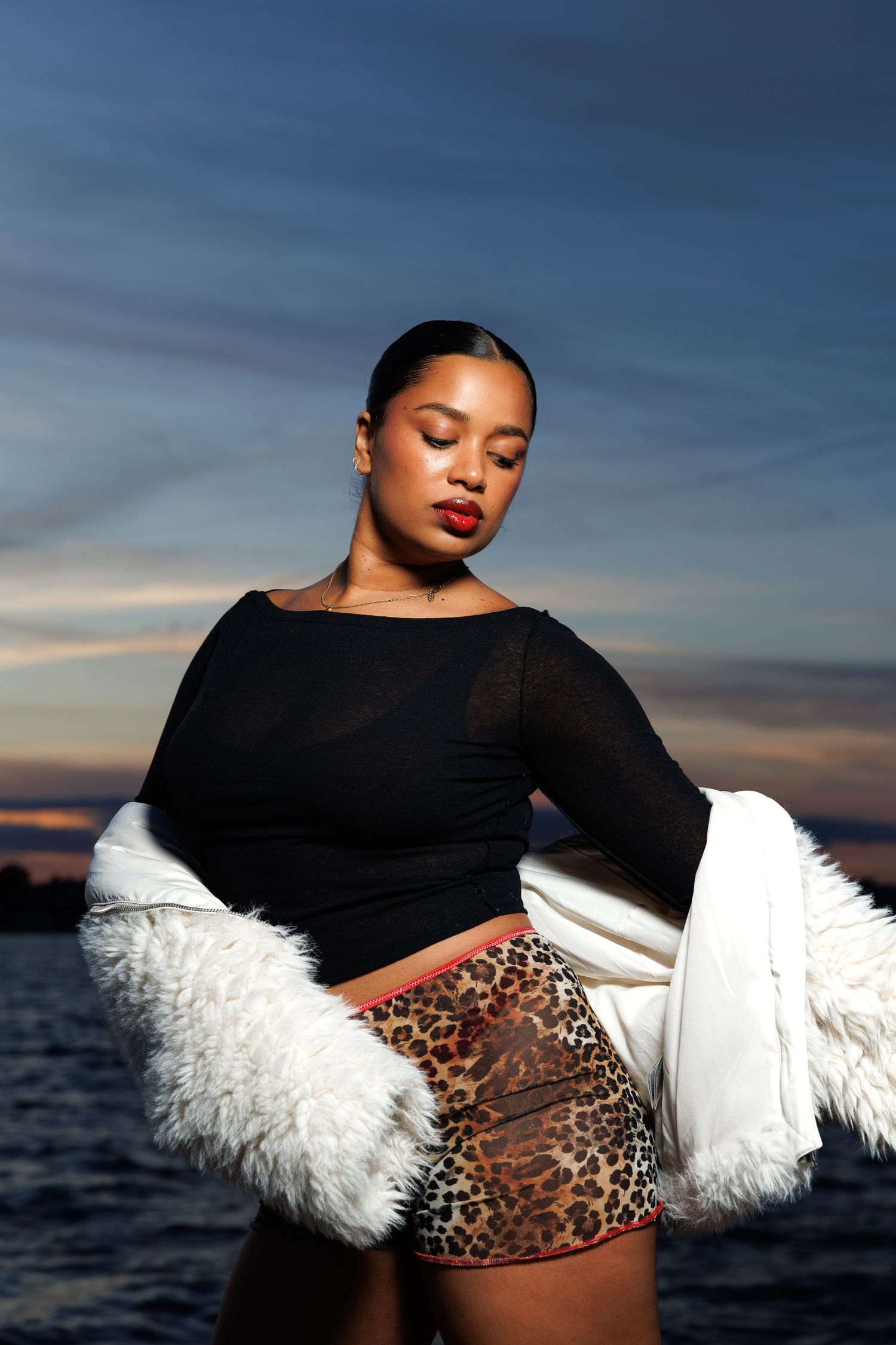 Woman in black top, leopard skirt, and white fur coat posing outdoors at sunset near water.