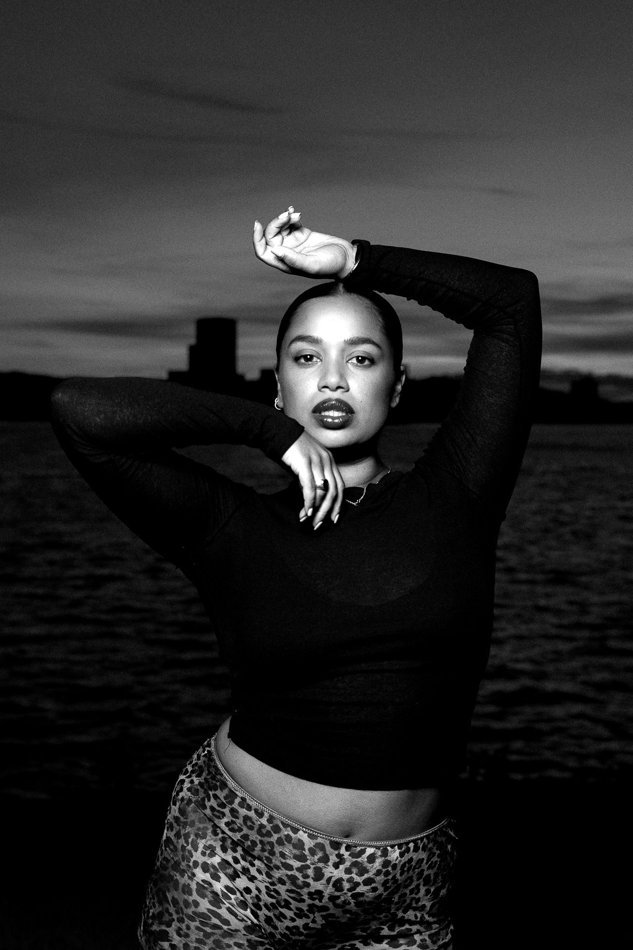 Black and white portrait of a woman in crop top and leopard print pants posing outdoors near water at dusk.