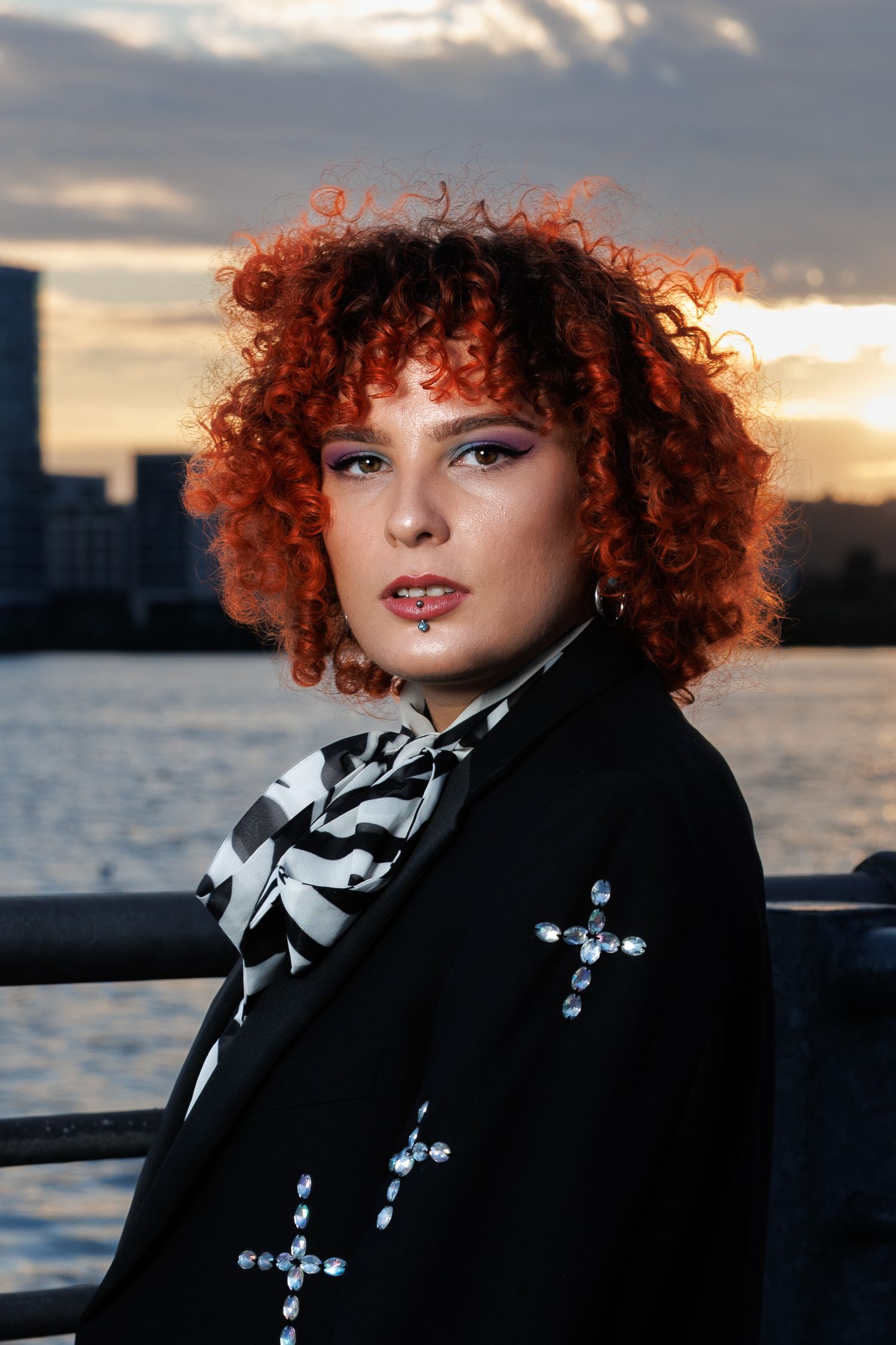 Woman with curly red hair wearing black blazer with cross embellishments and striped scarf at sunset waterfront.