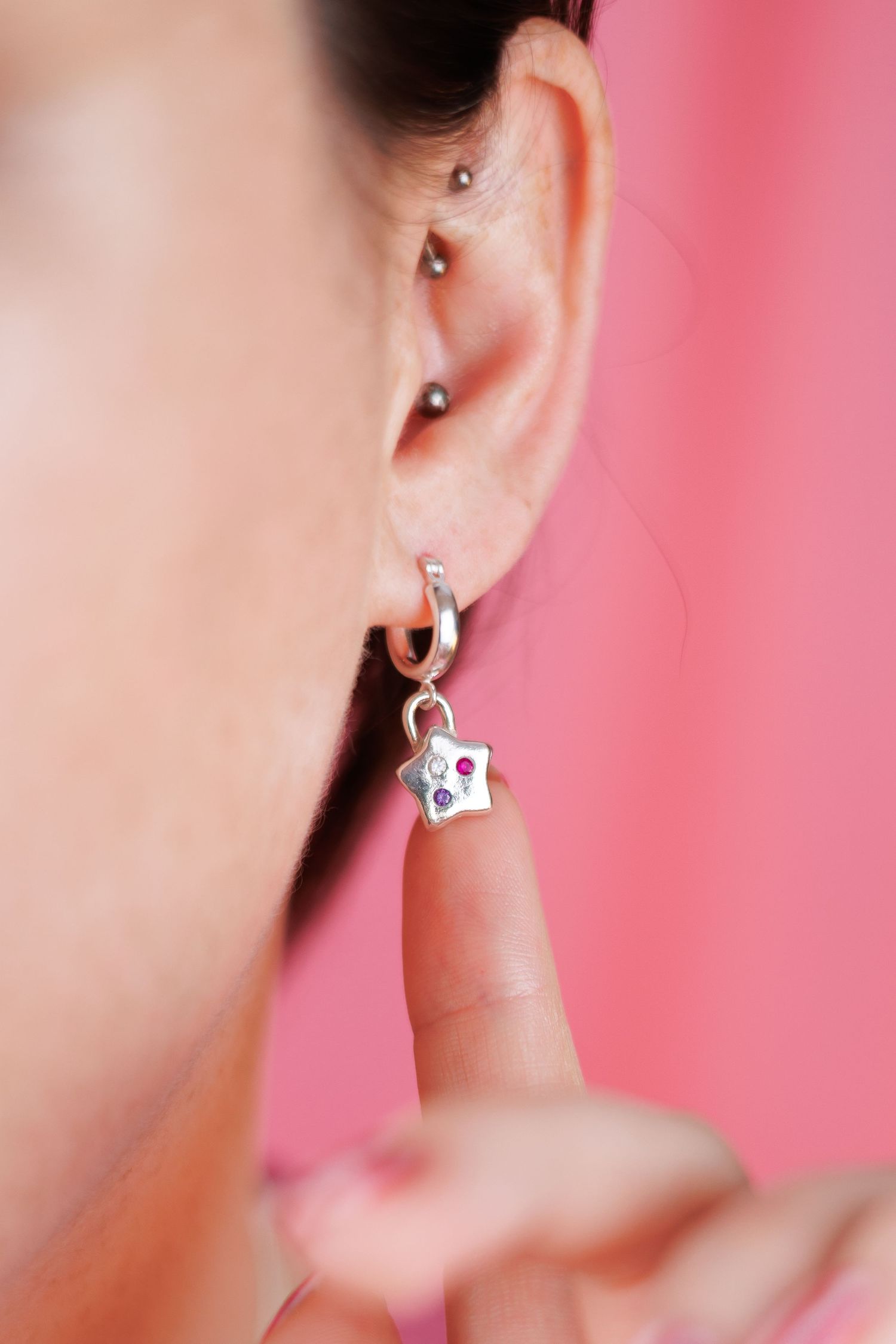 Close-up of a womans ear featuring a silver star charm earring with colorful gemstones on pink background.