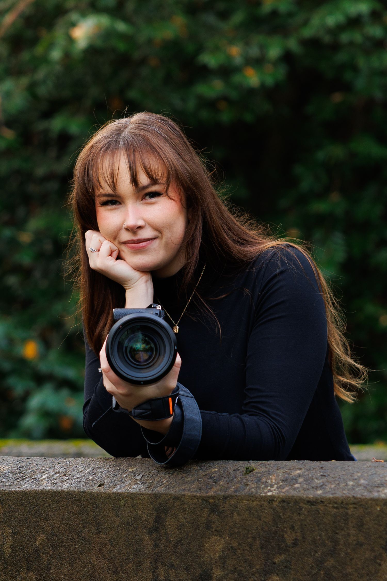 Young woman with long brown hair holding a professional camera while leaning on a stone wall.