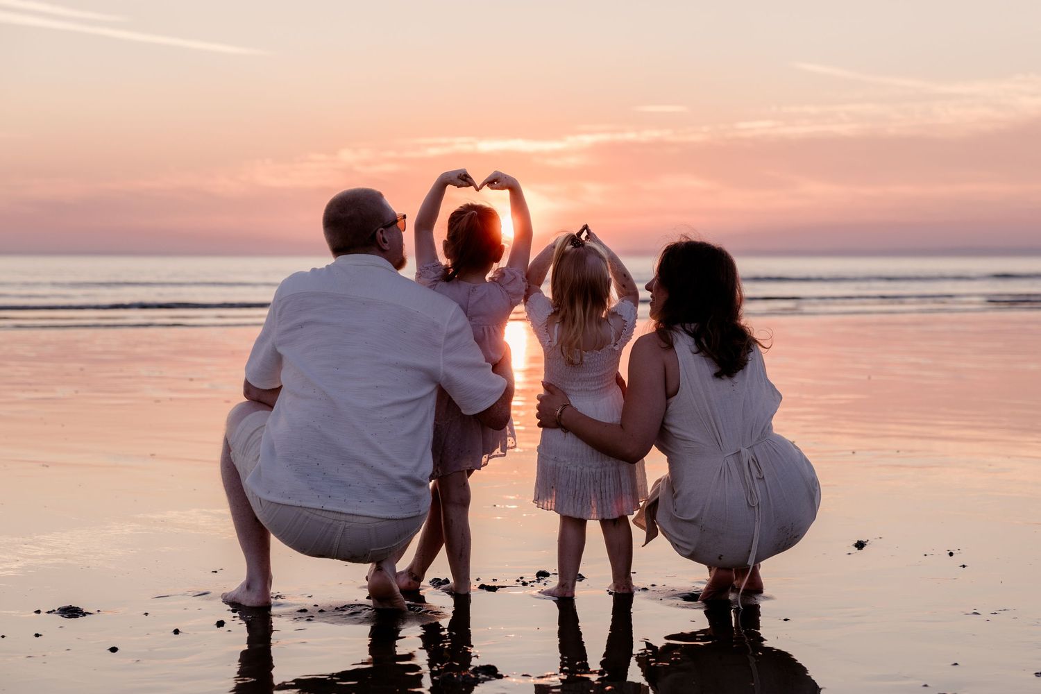 Family of four in white outfits forming heart shapes with hands at sunset on a beach.