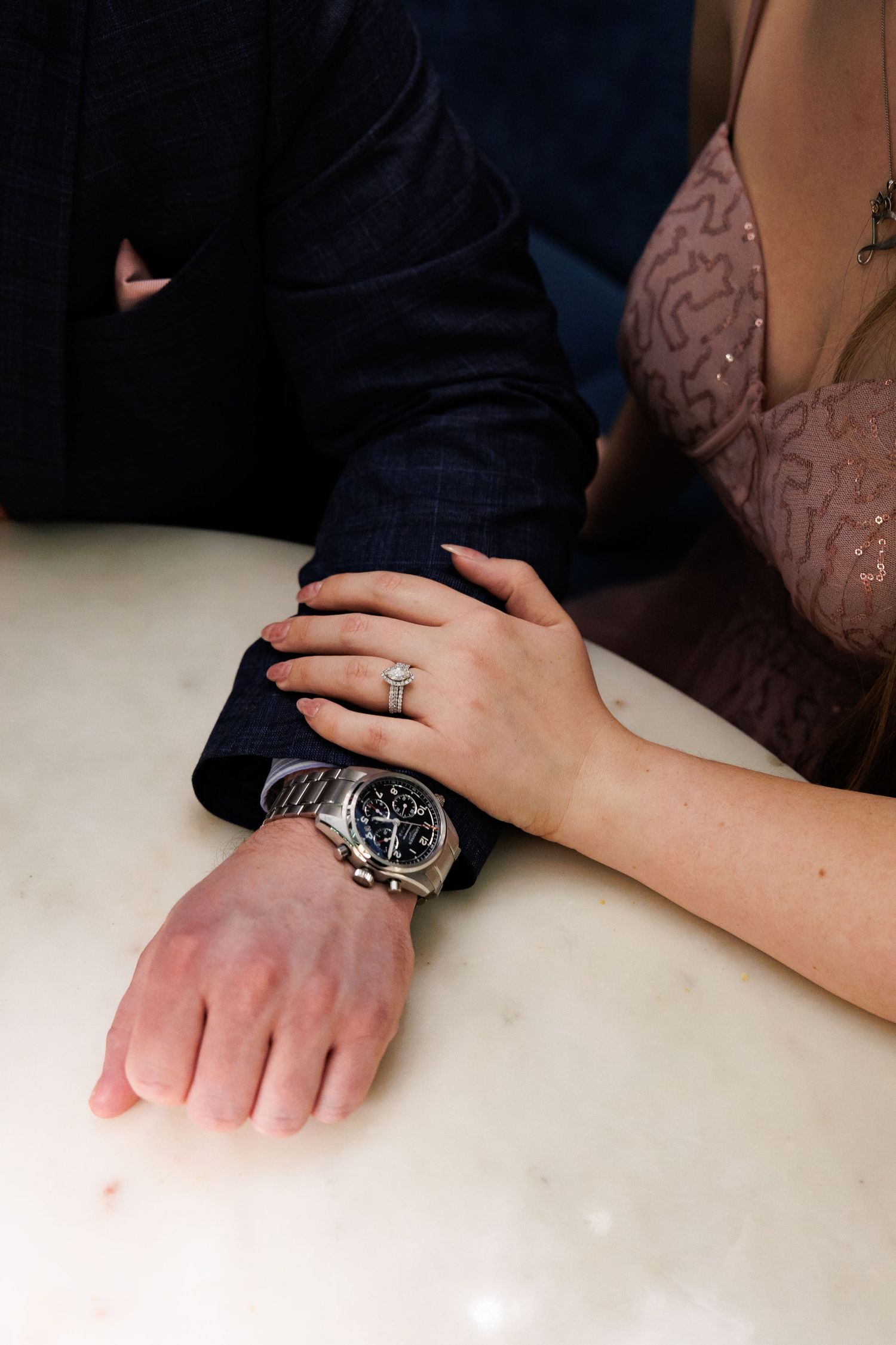 Close-up of a couple's hands on a marble surface, woman wearing diamond engagement ring, man wearing silver chronograph watch.
