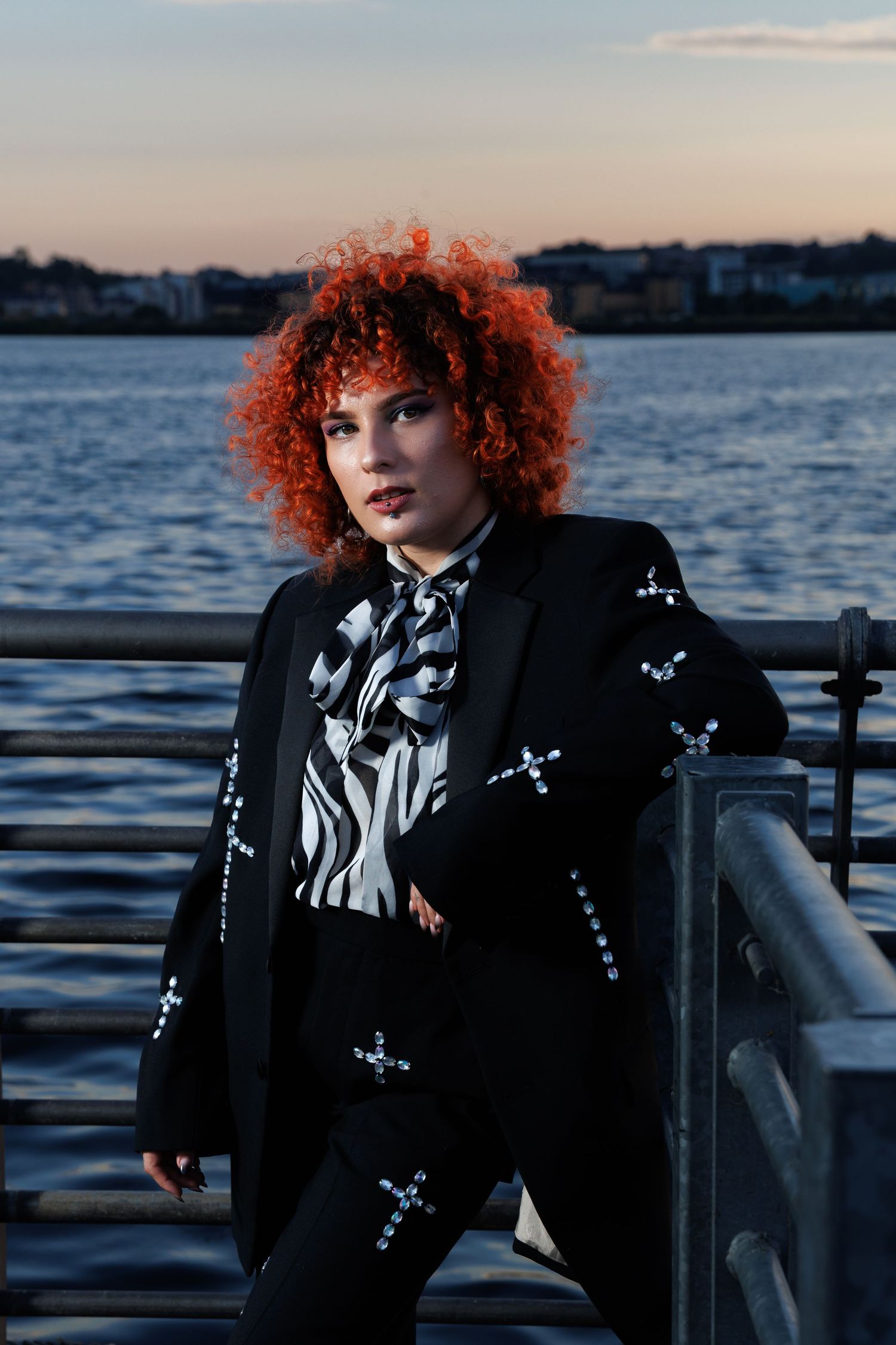 Woman with red curly hair in black embellished suit poses at waterfront railing at dusk.
