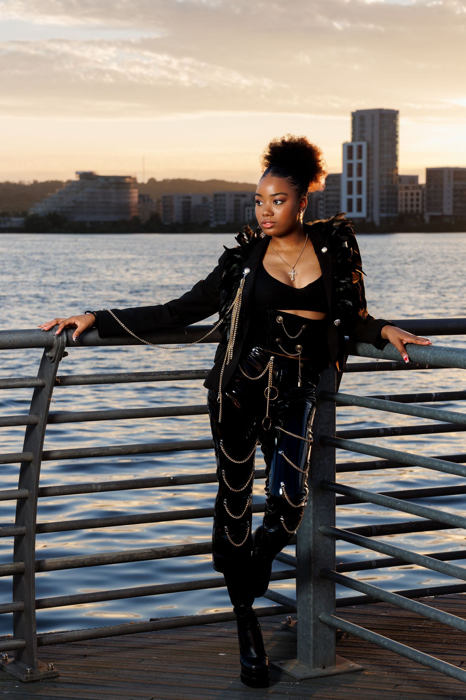 Woman in black studded jacket with gold chains poses at waterfront railing during golden hour sunset.