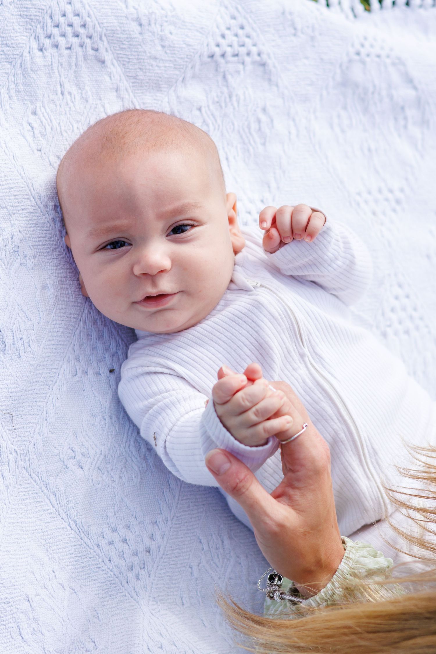 Newborn baby in white onesie lying on white blanket, gently holding an adult's finger.