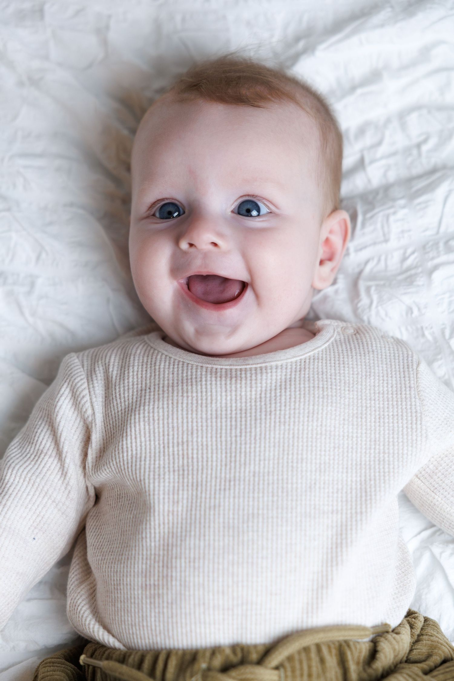Happy blue-eyed baby lying on white bedding, smiling in a cream ribbed top and olive pants.