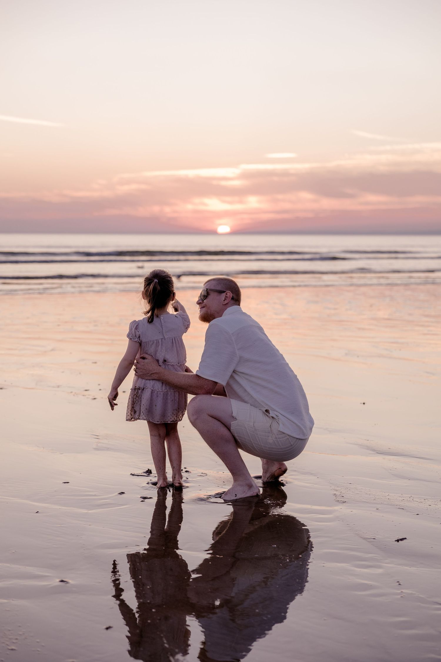 Father crouching beside young daughter on a wet beach at sunset, sharing a tender moment together.