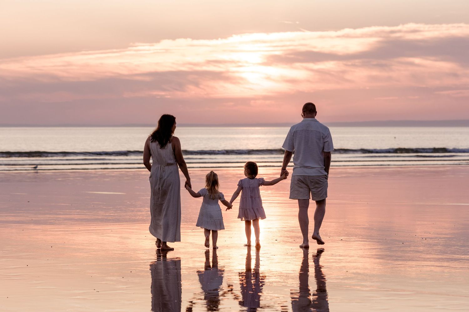 Family of four holding hands on a reflective beach at sunset, wearing white outfits with two young girls between parents.