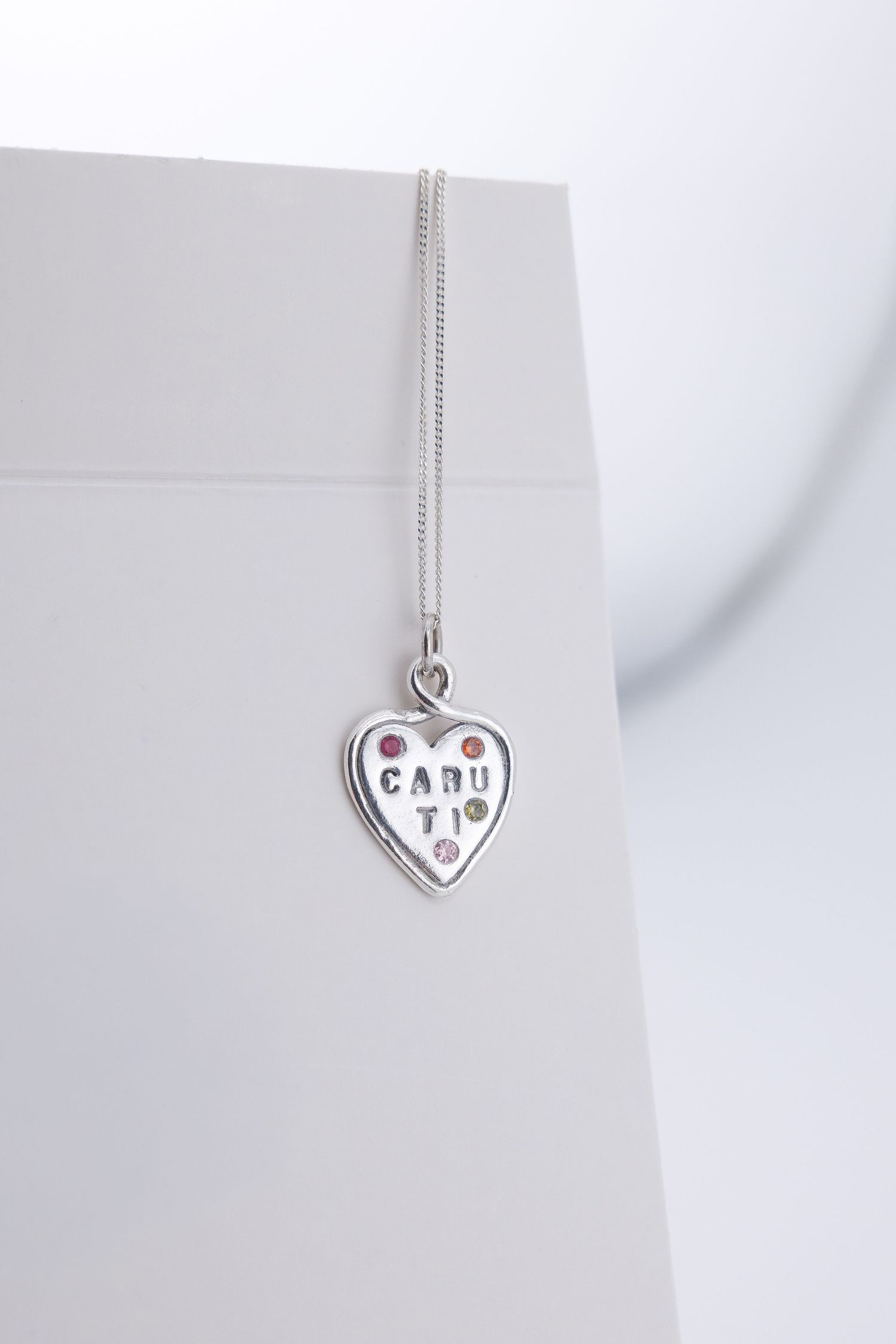 Silver heart-shaped pendant necklace engraved with CARU TI and colorful gemstone accents.