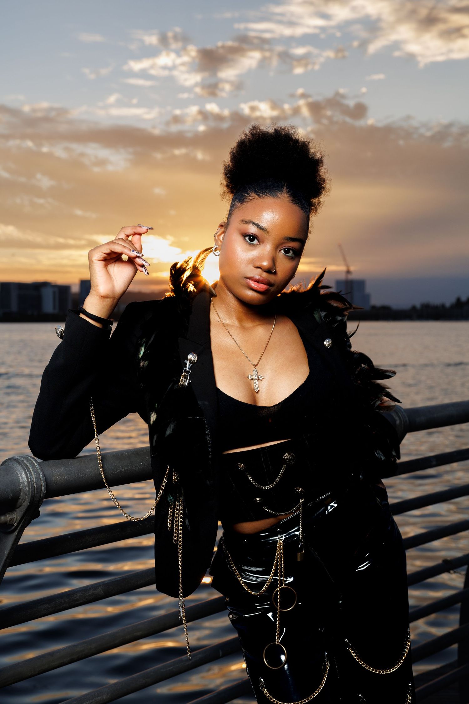 Stylish young Black woman in black feathered moto jacket with gold chains poses at pier during sunset.