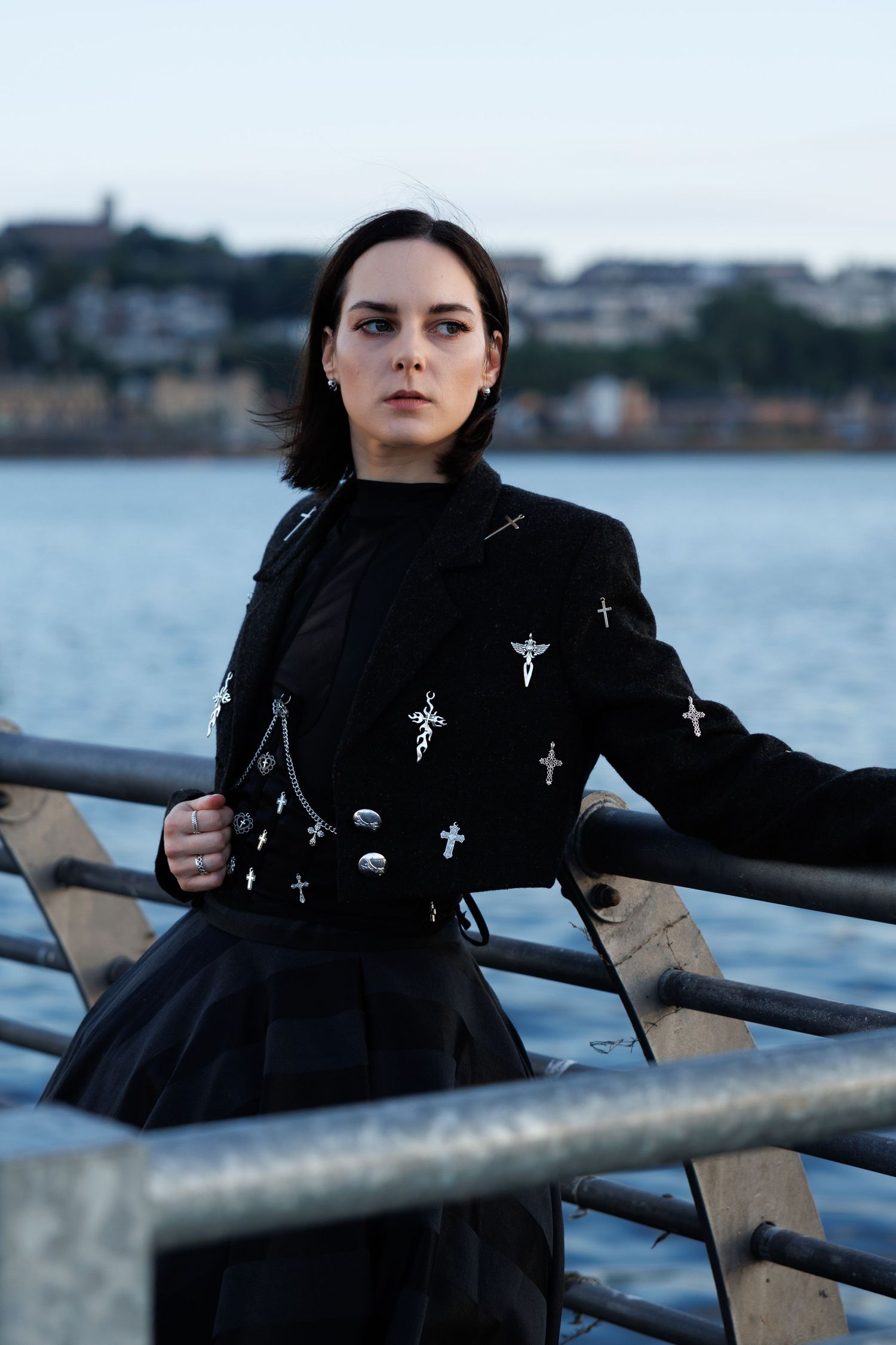 Woman in black cross-embellished jacket poses by waterfront railing at dusk.