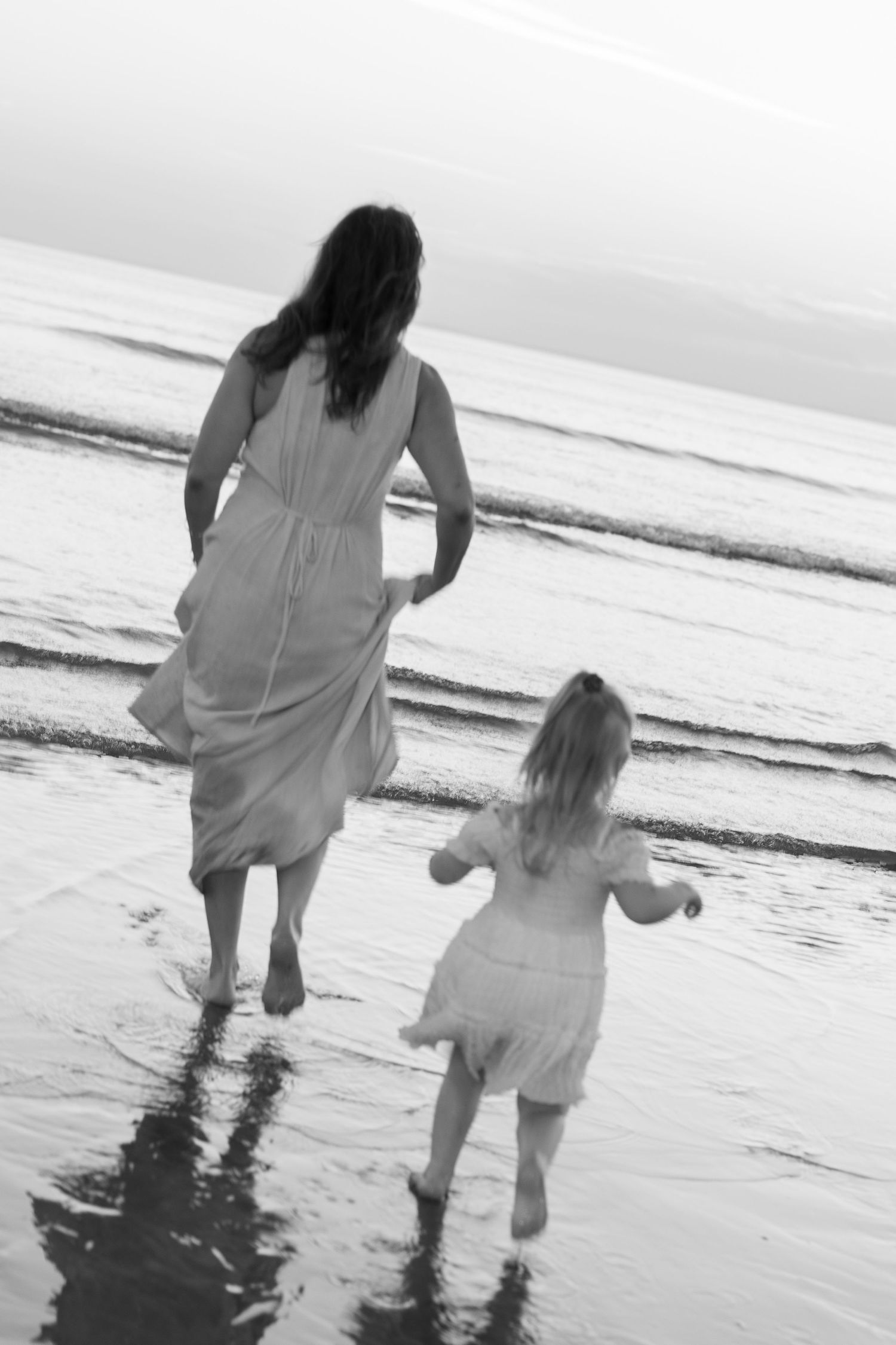 Black and white photo of mother and daughter running barefoot into ocean waves on beach.