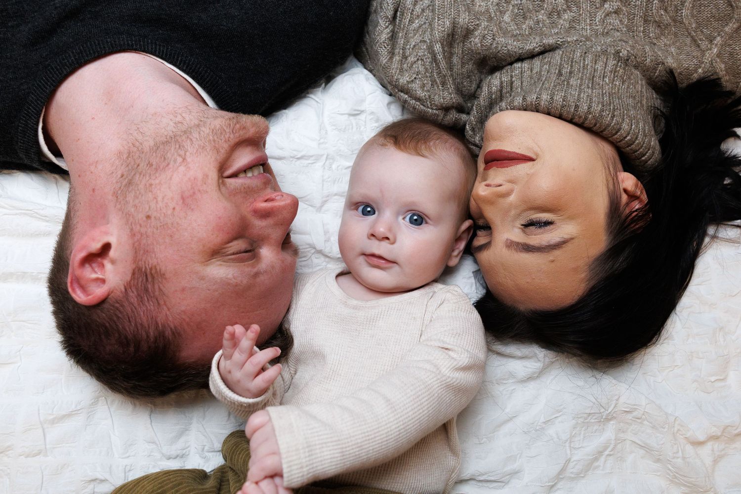 Two parents lying on a white bed with their blue-eyed baby between them smiling at the camera.