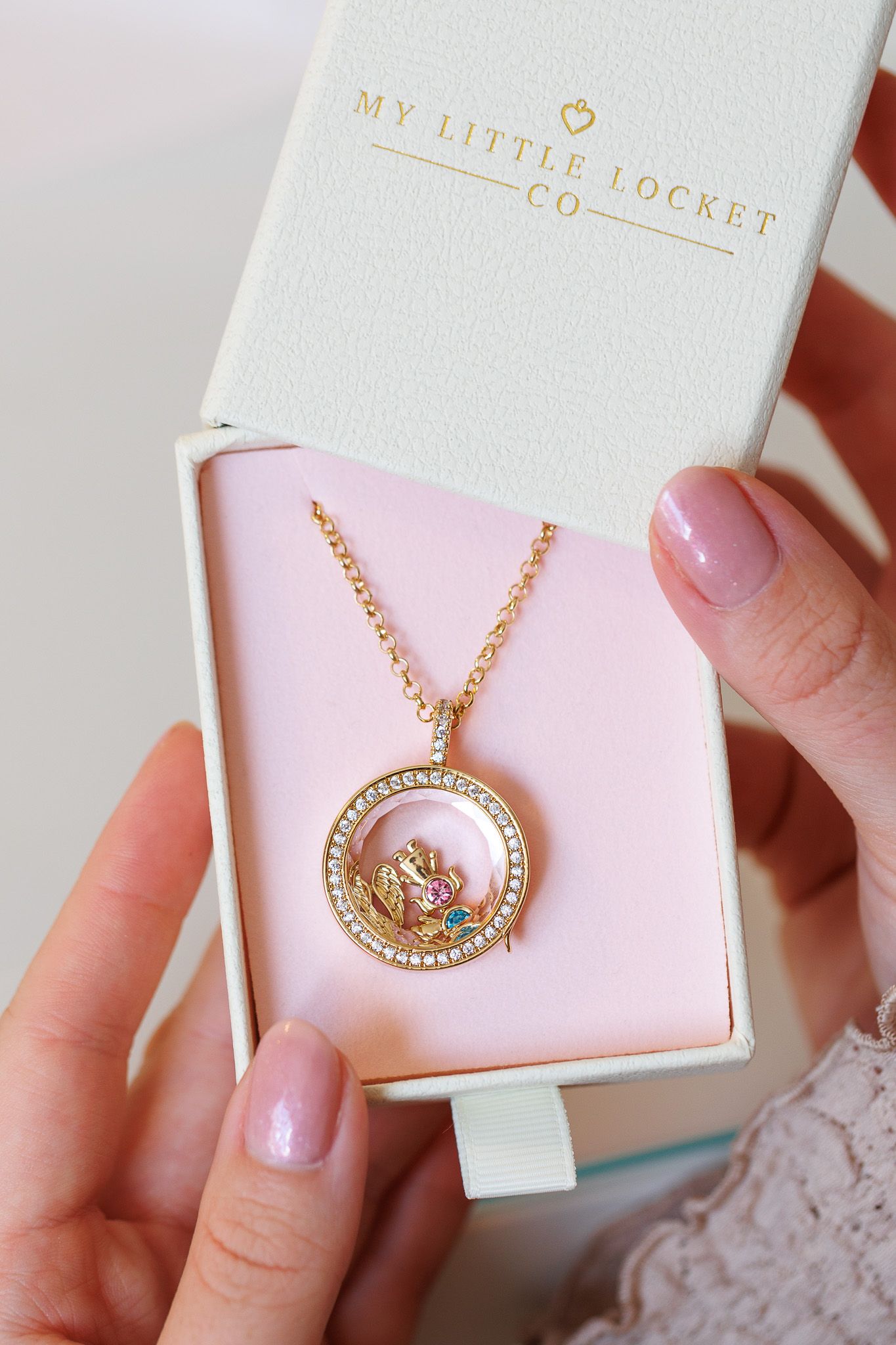 Hands holding a My Little Locket Co. gold circular locket necklace with crystals in a pink gift box.