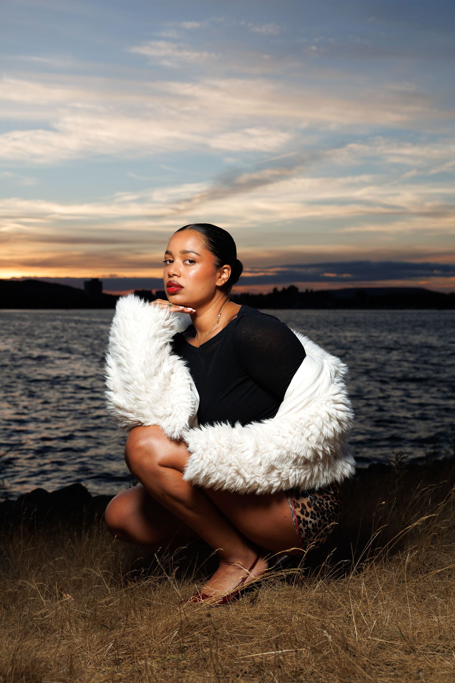 Woman in white fur coat and leopard skirt poses by a lake at sunset in a crouching position.