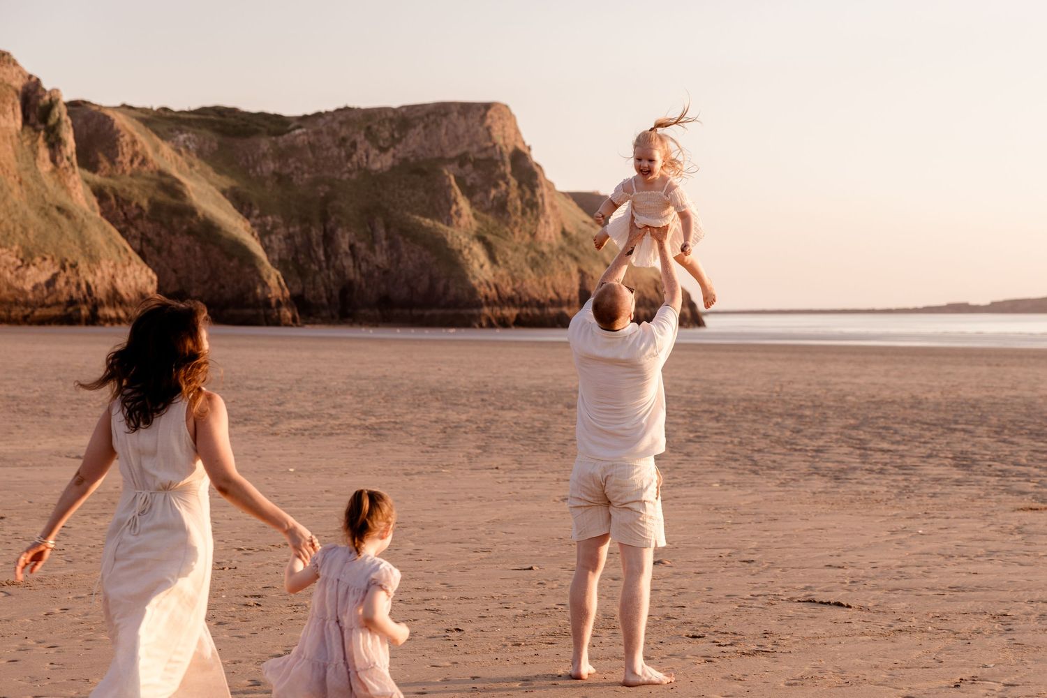 Family in white outfits playing on a sandy beach at sunset, father tossing laughing child in the air near coastal cliffs.