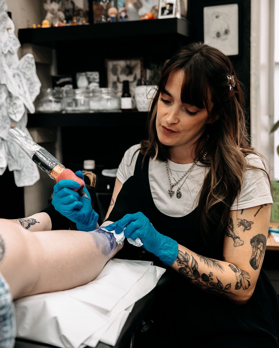 Female tattoo artist with sleeve tattoos carefully tattooing a clients leg in studio.