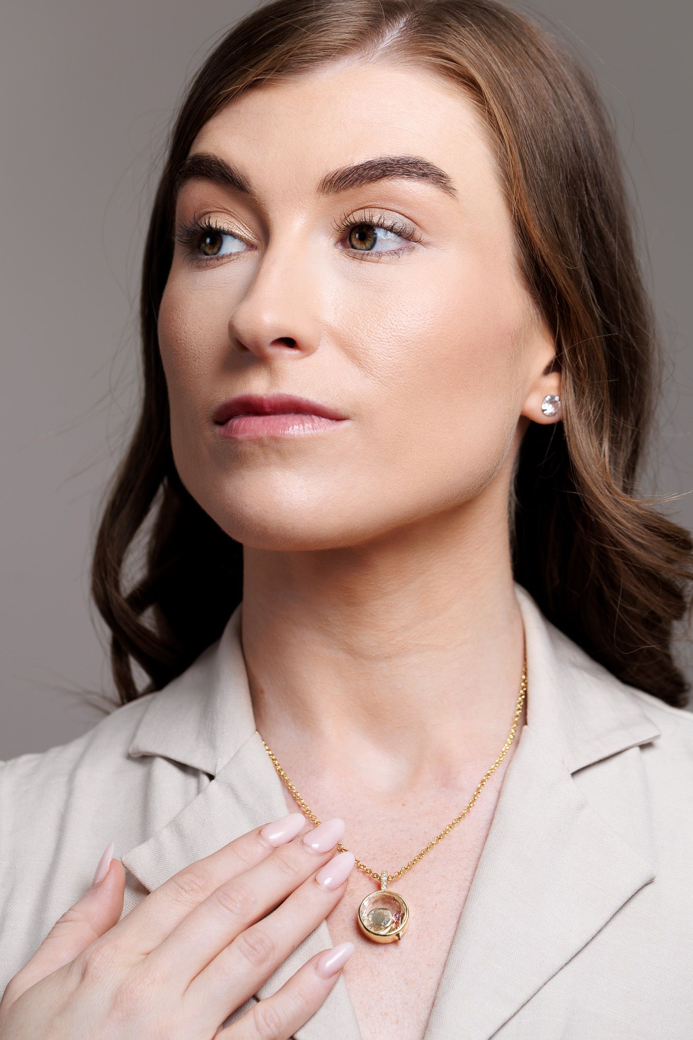 Woman wearing gold chain necklace with round pendant and stud earrings, dressed in beige blazer against gray background.