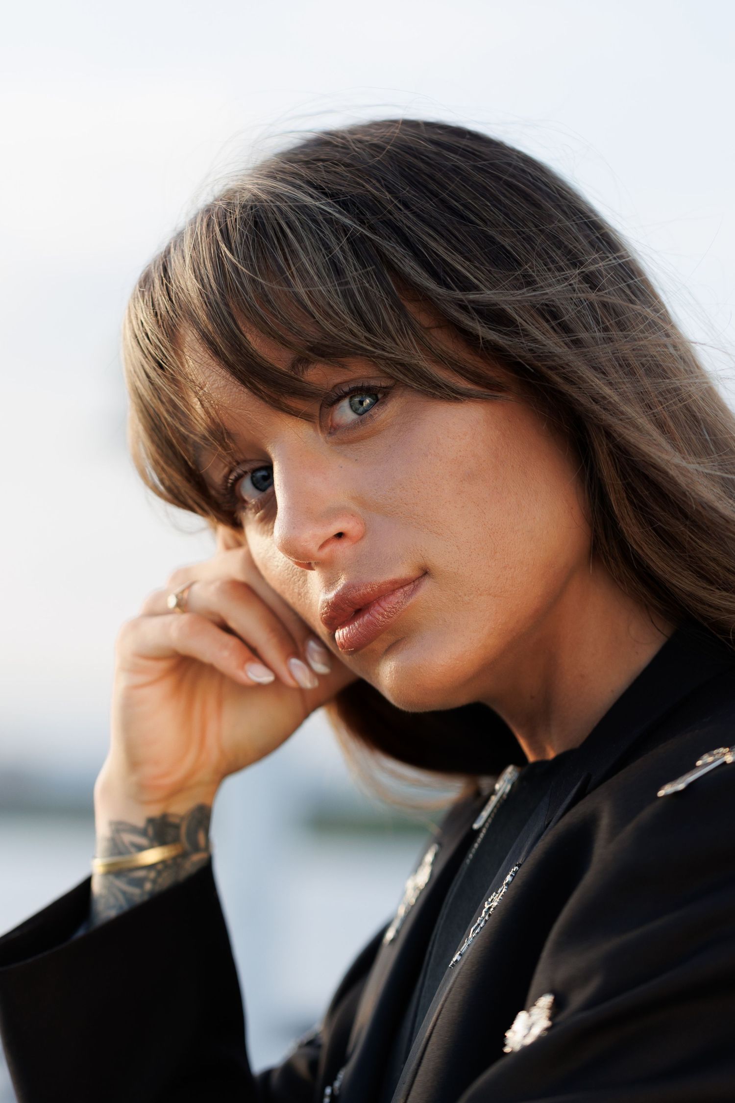 A young woman with brown hair and bangs rests her chin on her hand, wearing a black jacket with tattoos visible.