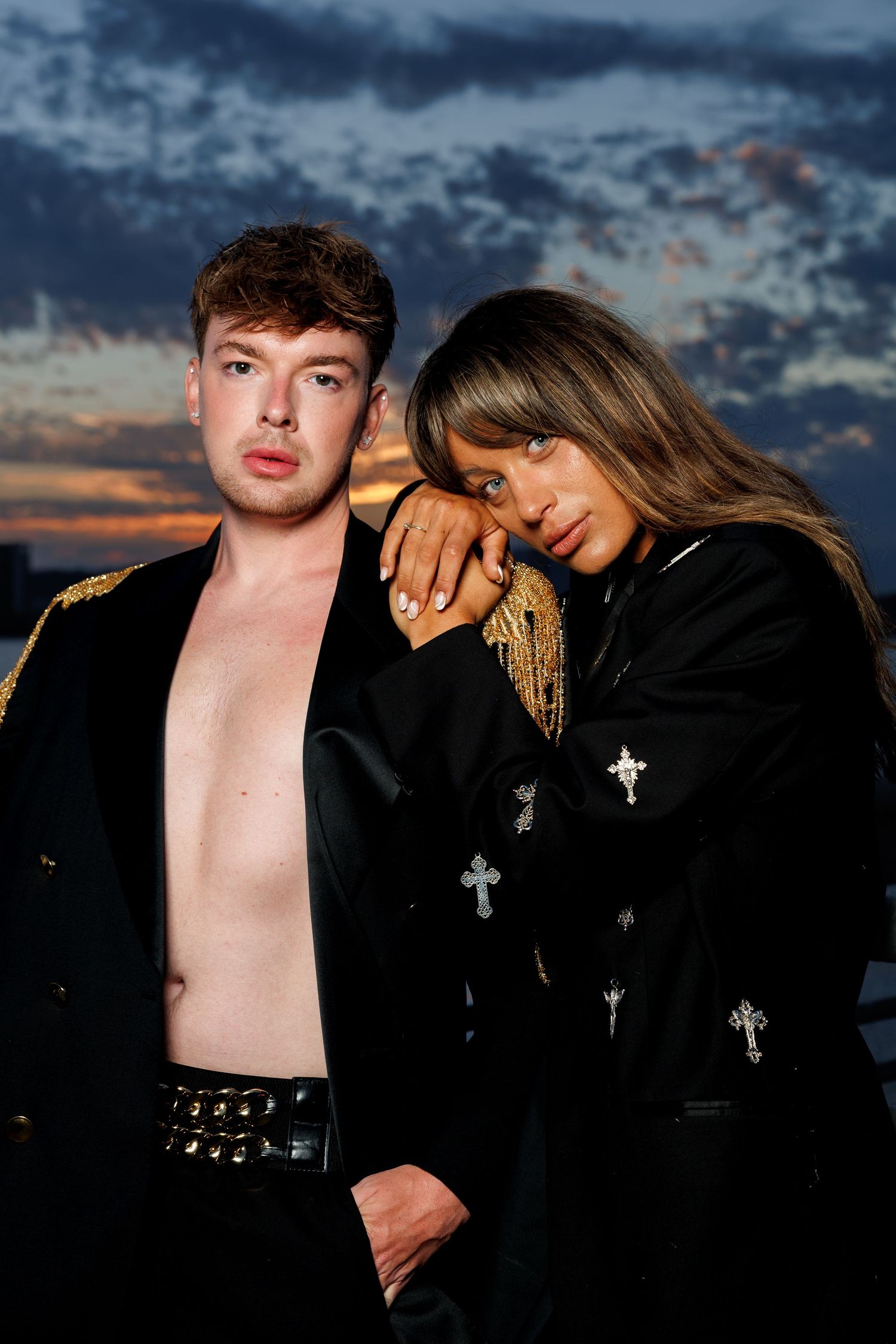 A man and woman pose dramatically at sunset, wearing black jackets with cross embellishments and gold epaulettes.
