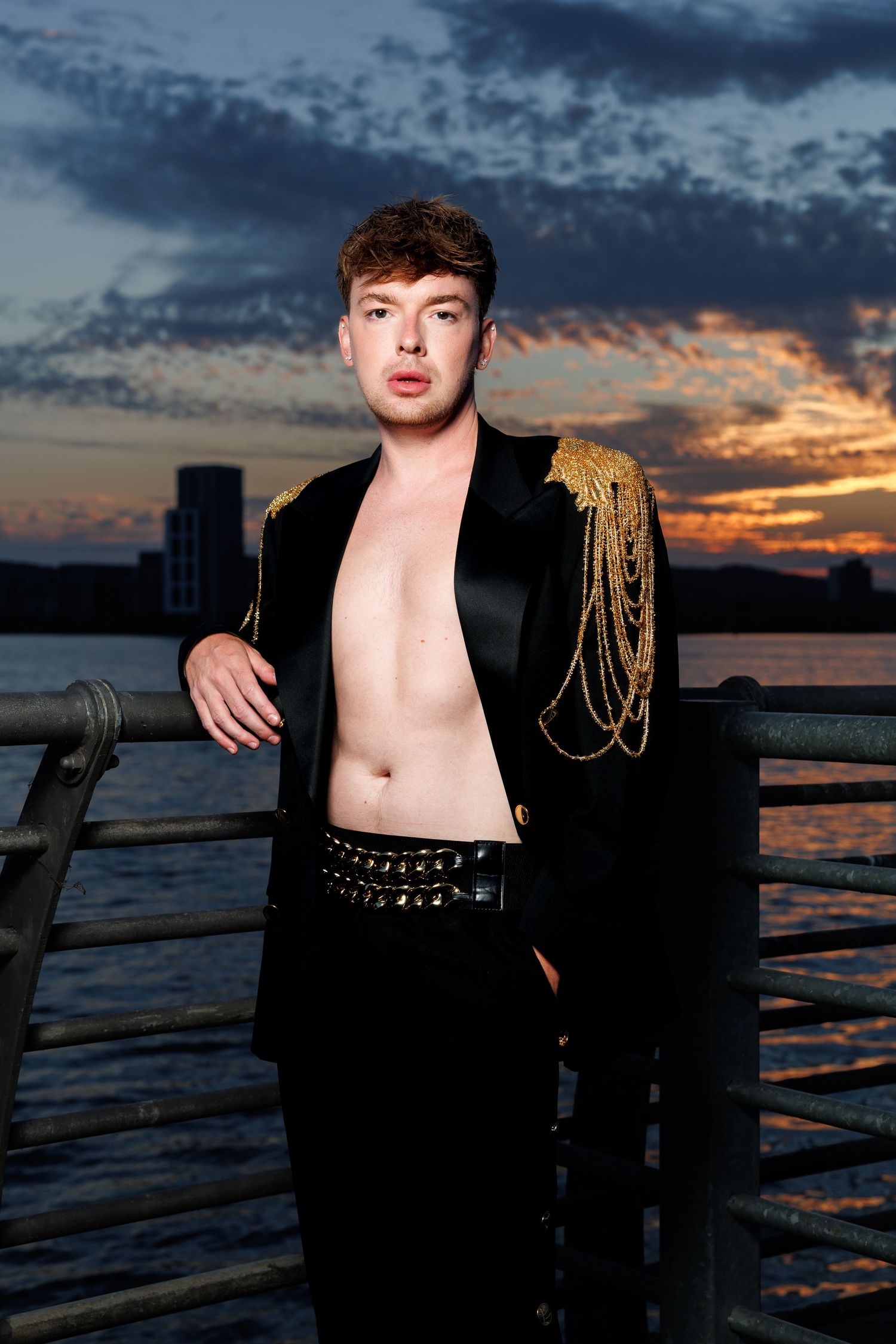 Young man in black jacket with gold chains poses shirtless on pier at dramatic sunset.