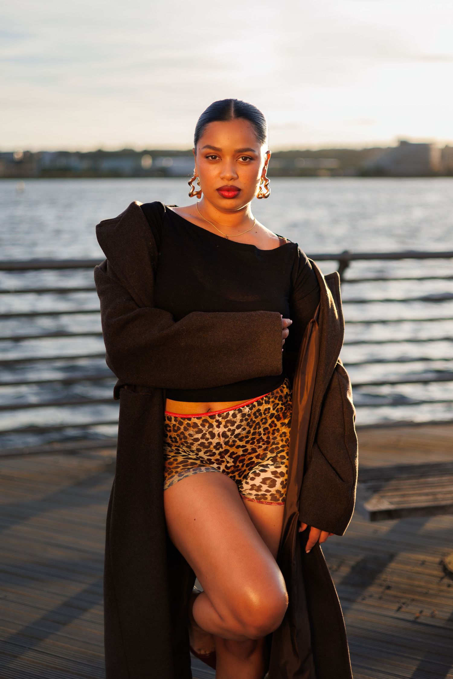 Woman in leopard print shorts and long black coat posing on a waterfront dock at sunset.