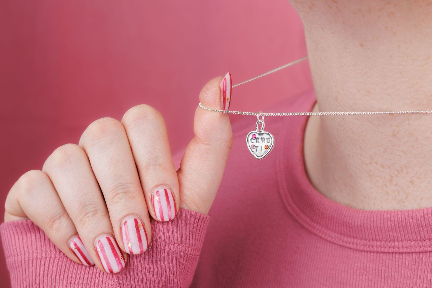 Woman in pink top displays heart pendant necklace with pink striped manicured nails on pink background.