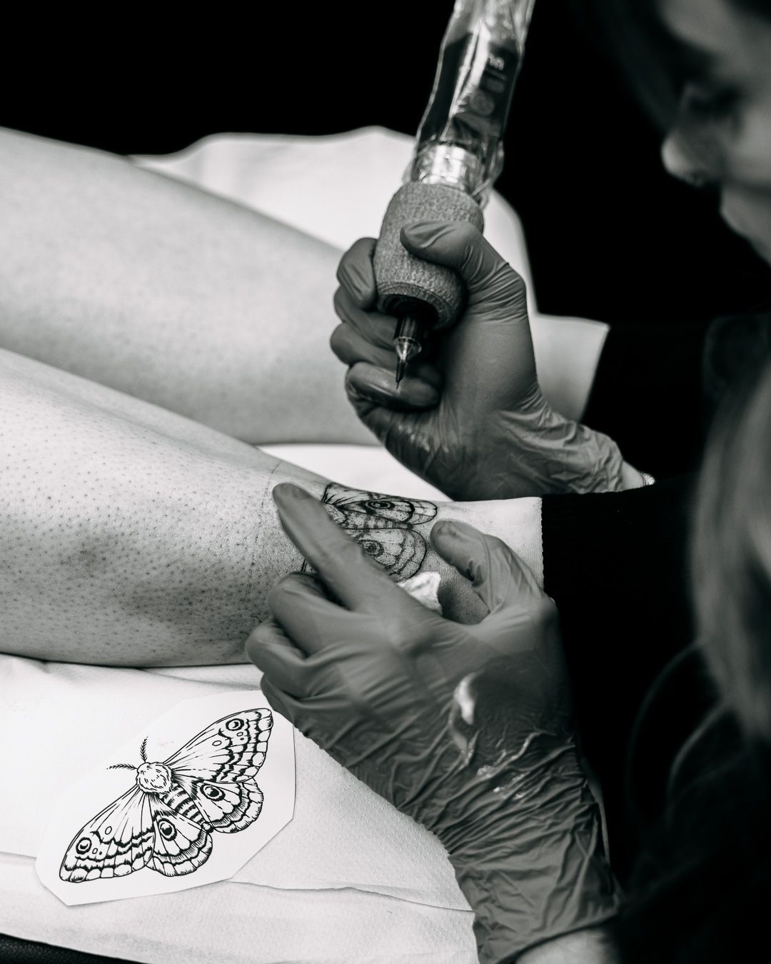 Black and white photo of a tattoo artist carefully inking a detailed moth tattoo on a clients leg.