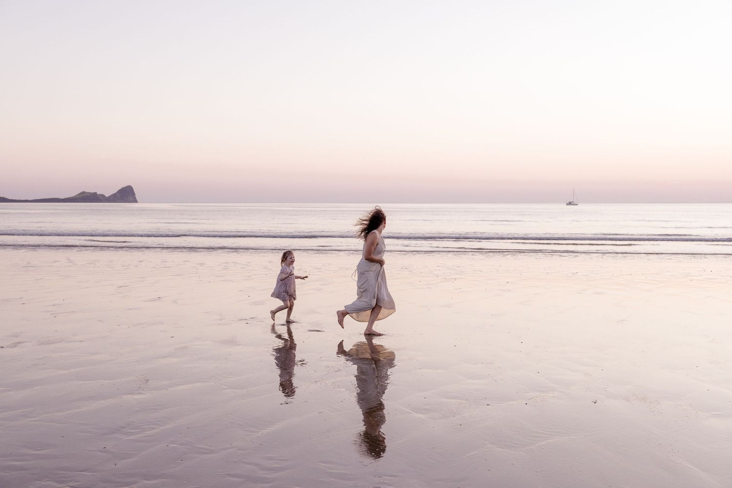 A mother and child run joyfully along a serene beach at sunset, their reflections visible on the wet sand.