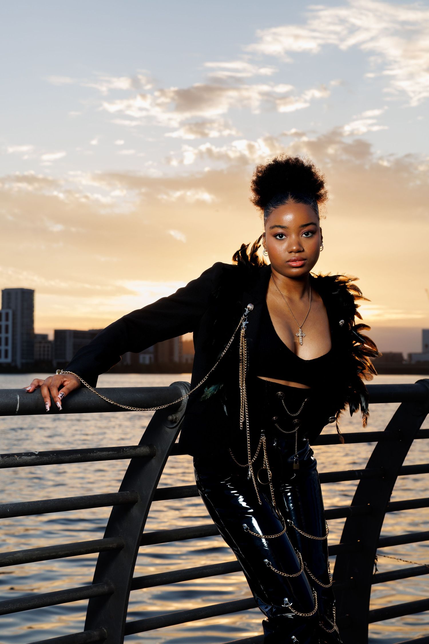 Young Black woman in black feathered jacket and chain pants poses at waterfront railing during golden sunset.
