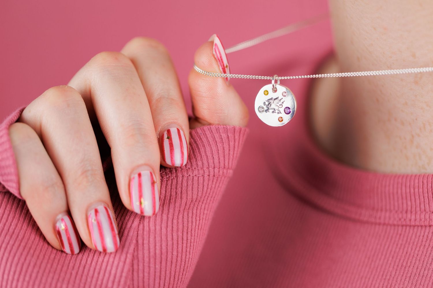 Close-up of hand with pink striped nails holding a silver circular charm necklace pendant.