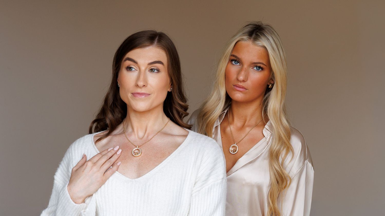 Two women in white silk tops modeling gold heart necklaces against a neutral background.