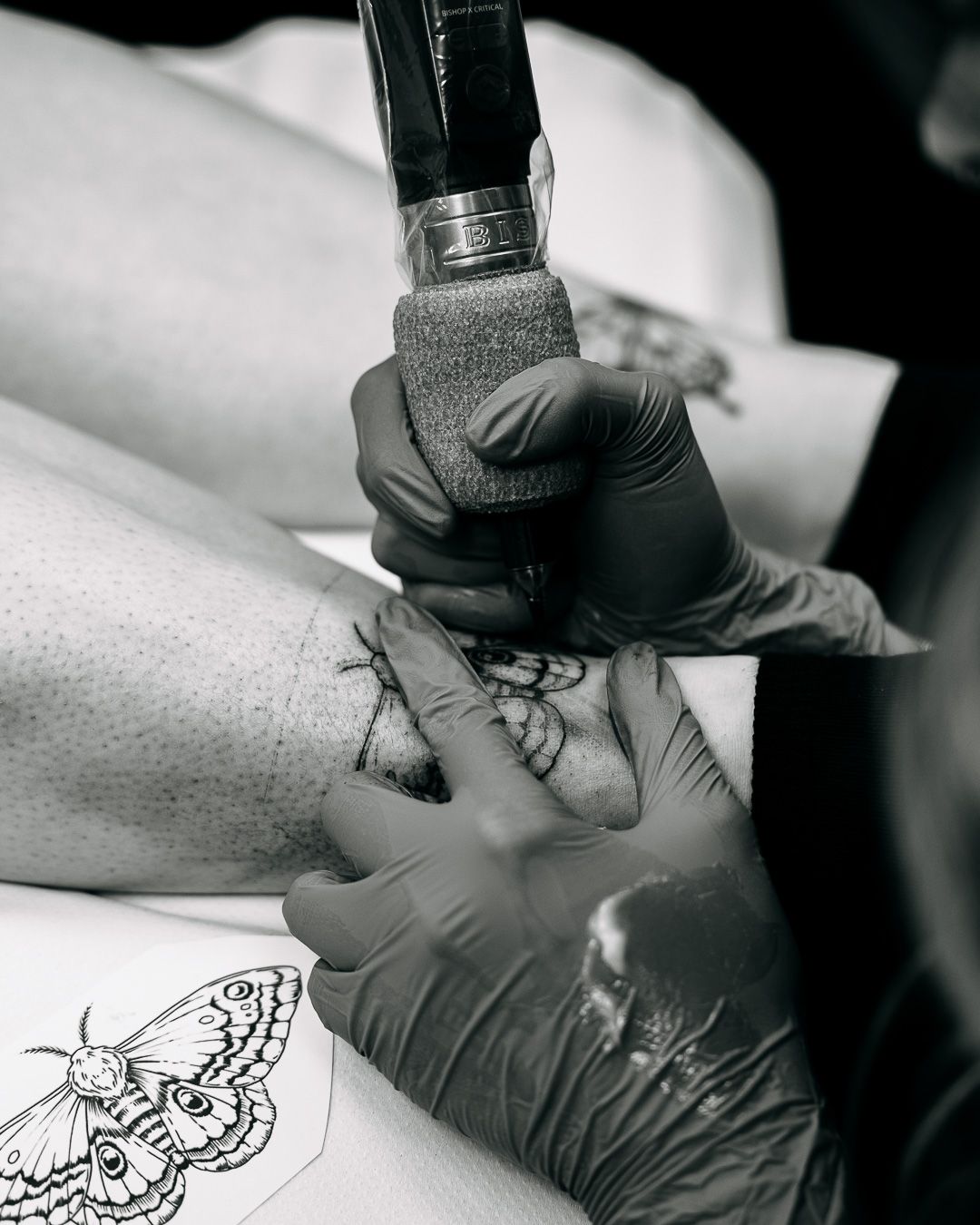 Black and white close-up of tattoo artist applying detailed moth design to client's leg using tattoo machine.