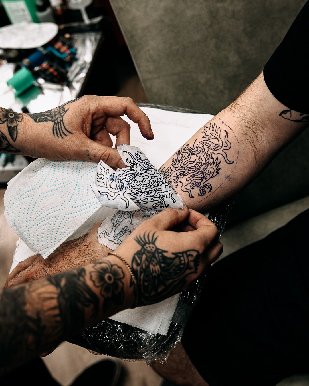 Tattoo artist placing stencil design on heavily tattooed clients forearm before inking.