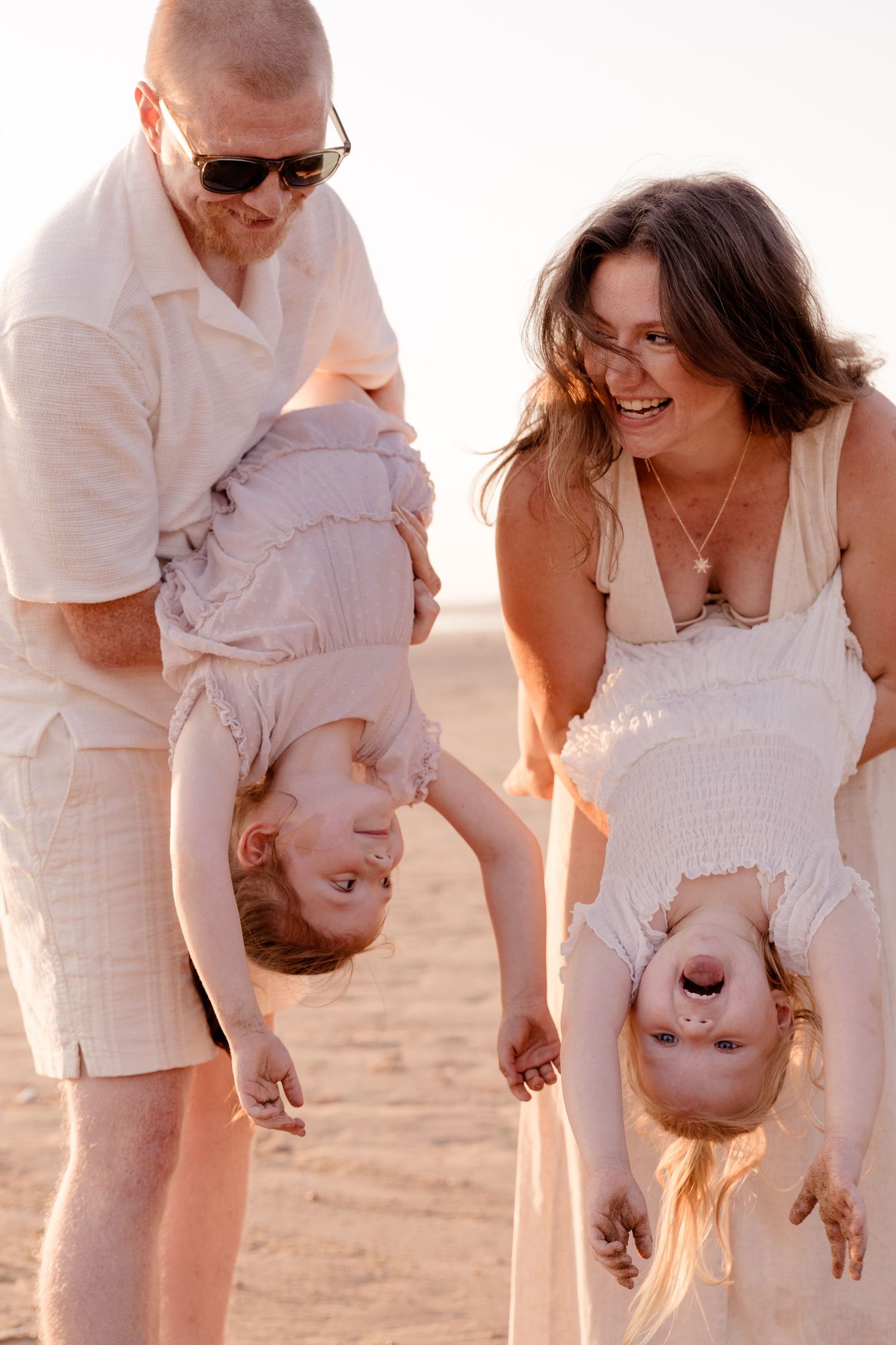 Happy family at beach, parents playfully holding two young children upside down, laughing in warm golden sunlight.