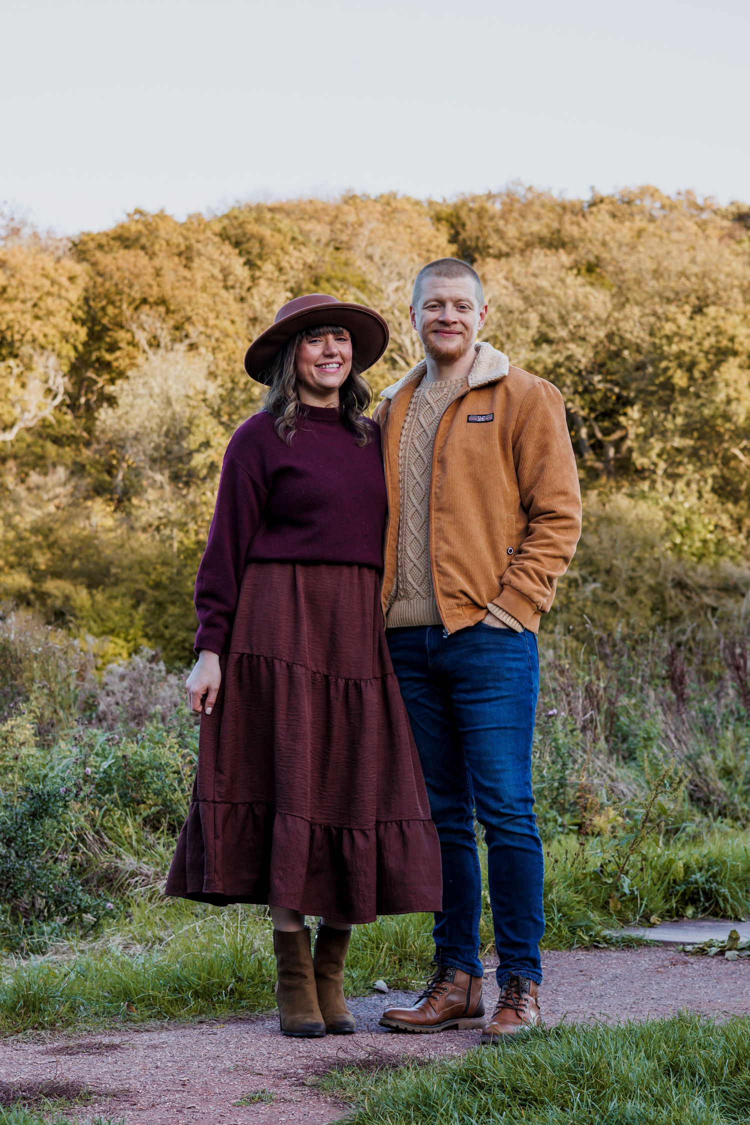 Couple posing outdoors in autumn, woman in burgundy dress and hat, man in tan jacket and jeans, fall foliage background.