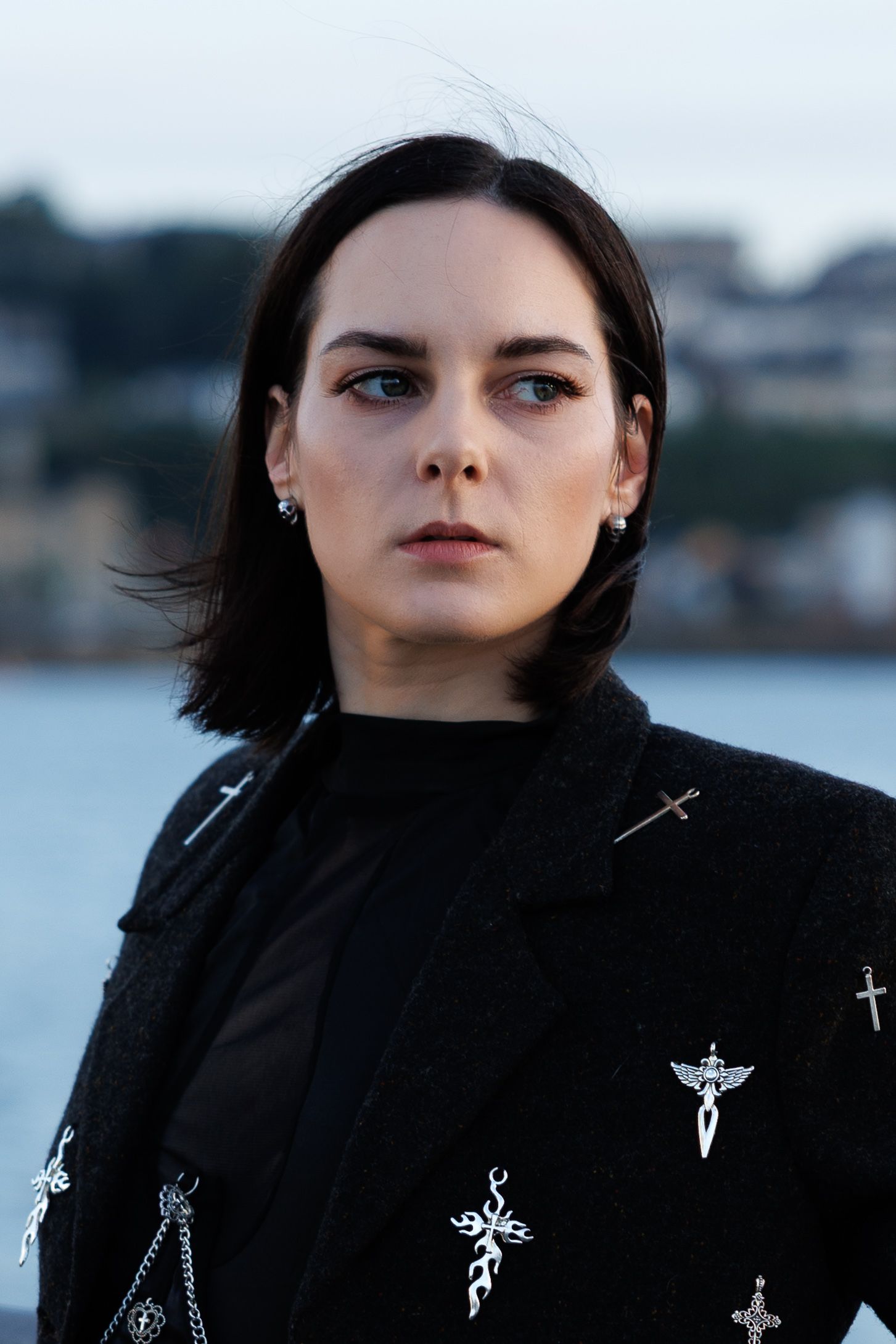 Young woman with dark hair wearing a black jacket adorned with silver cross pins, standing outdoors near water.