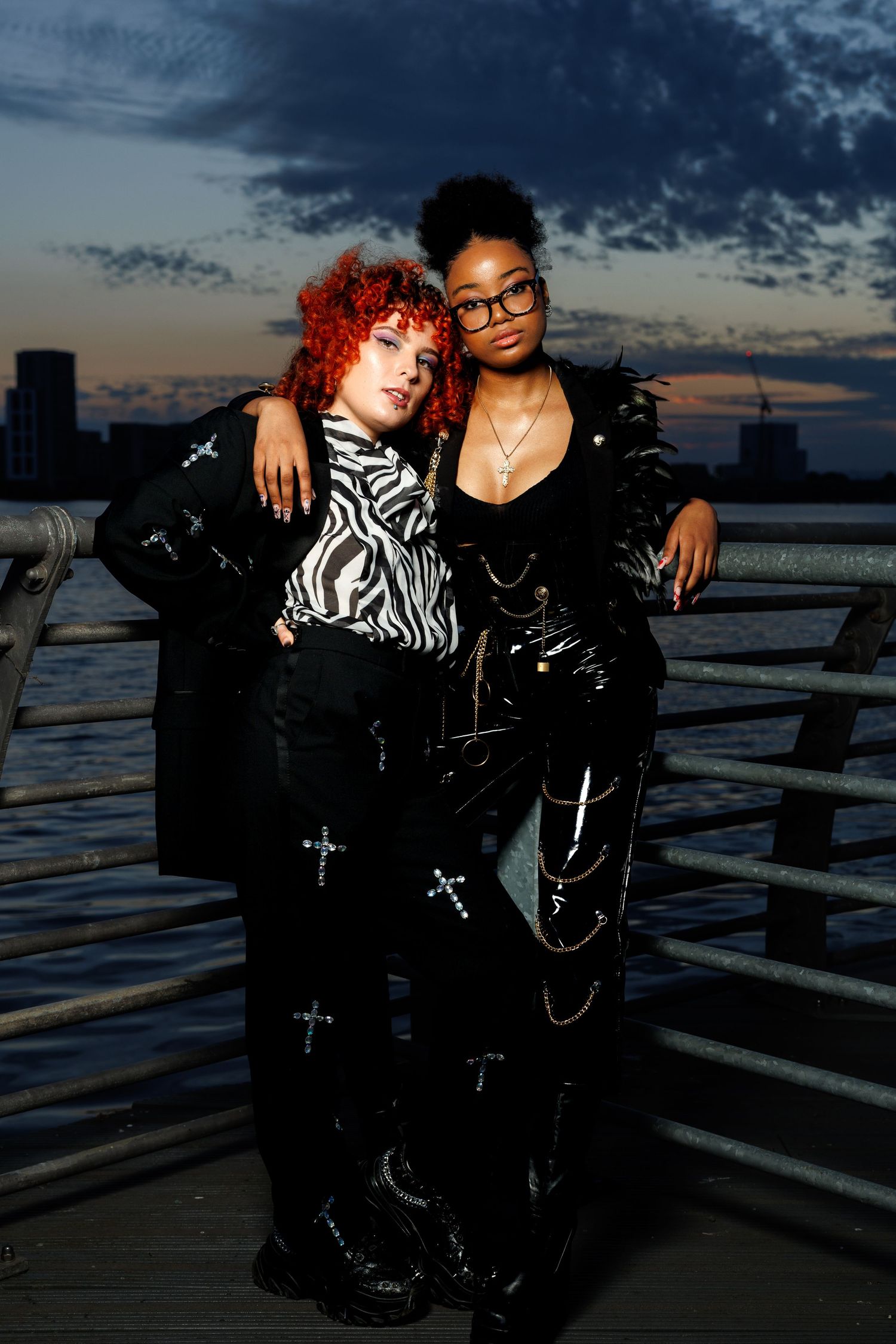 Two stylish women pose on a waterfront pier at dusk, wearing edgy black outfits with cross details and bold accessories.