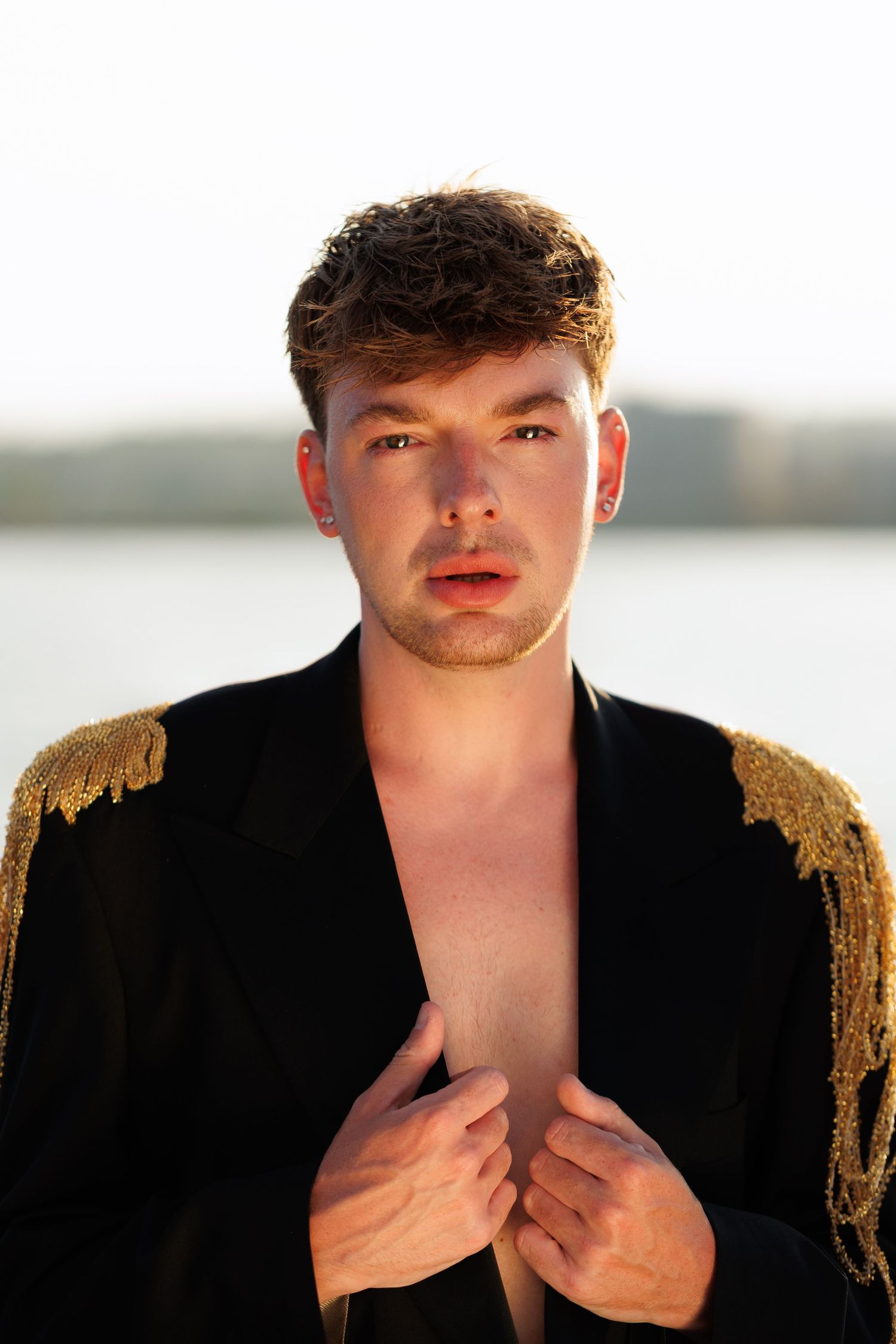 Young man in open black jacket with gold epaulettes posing outdoors near water in golden hour light.