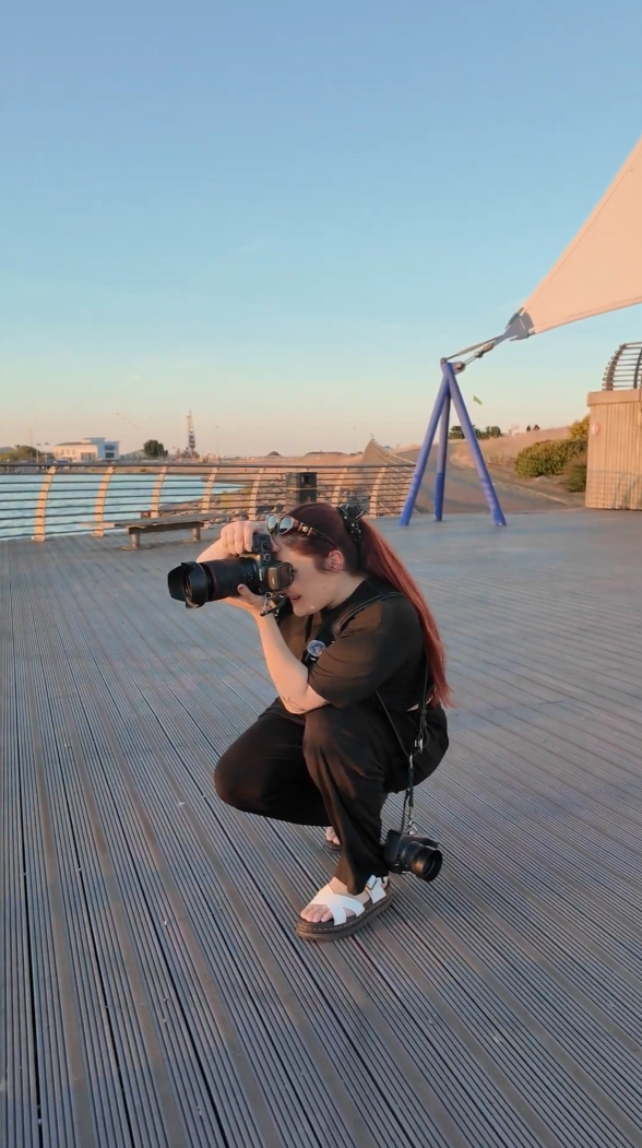 A woman with red hair crouches on a wooden boardwalk, photographing with a DSLR camera during golden hour sunset.