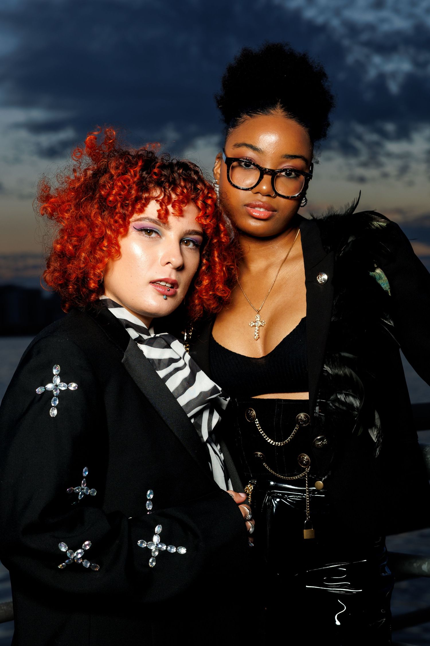 Two stylish women pose at dusk, one with red curly hair, one with glasses and chains, both in edgy black outfits.