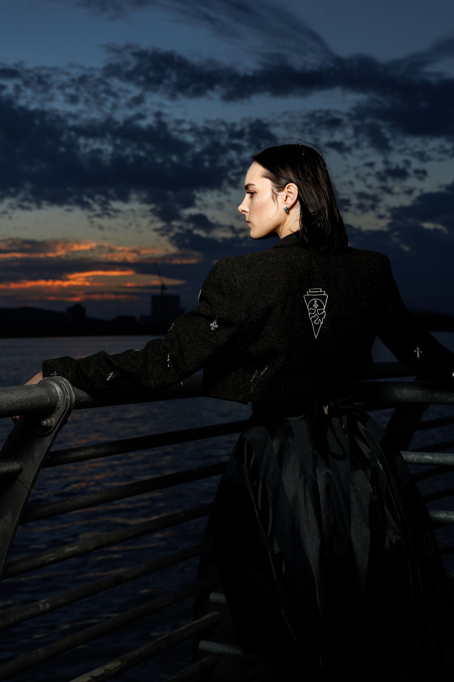 Person in black jacket and skirt leaning on railing at waterfront during dramatic sunset with dark clouds.