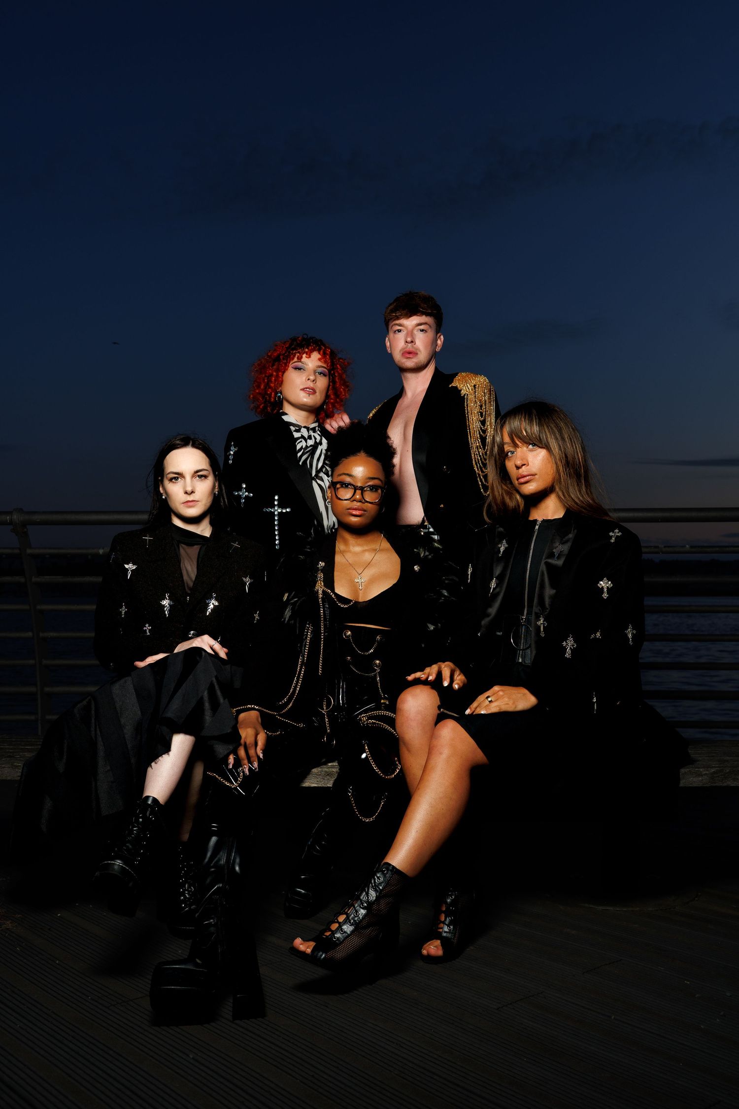 Five diverse young adults in dark gothic fashion pose together outdoors at dusk near a railing.