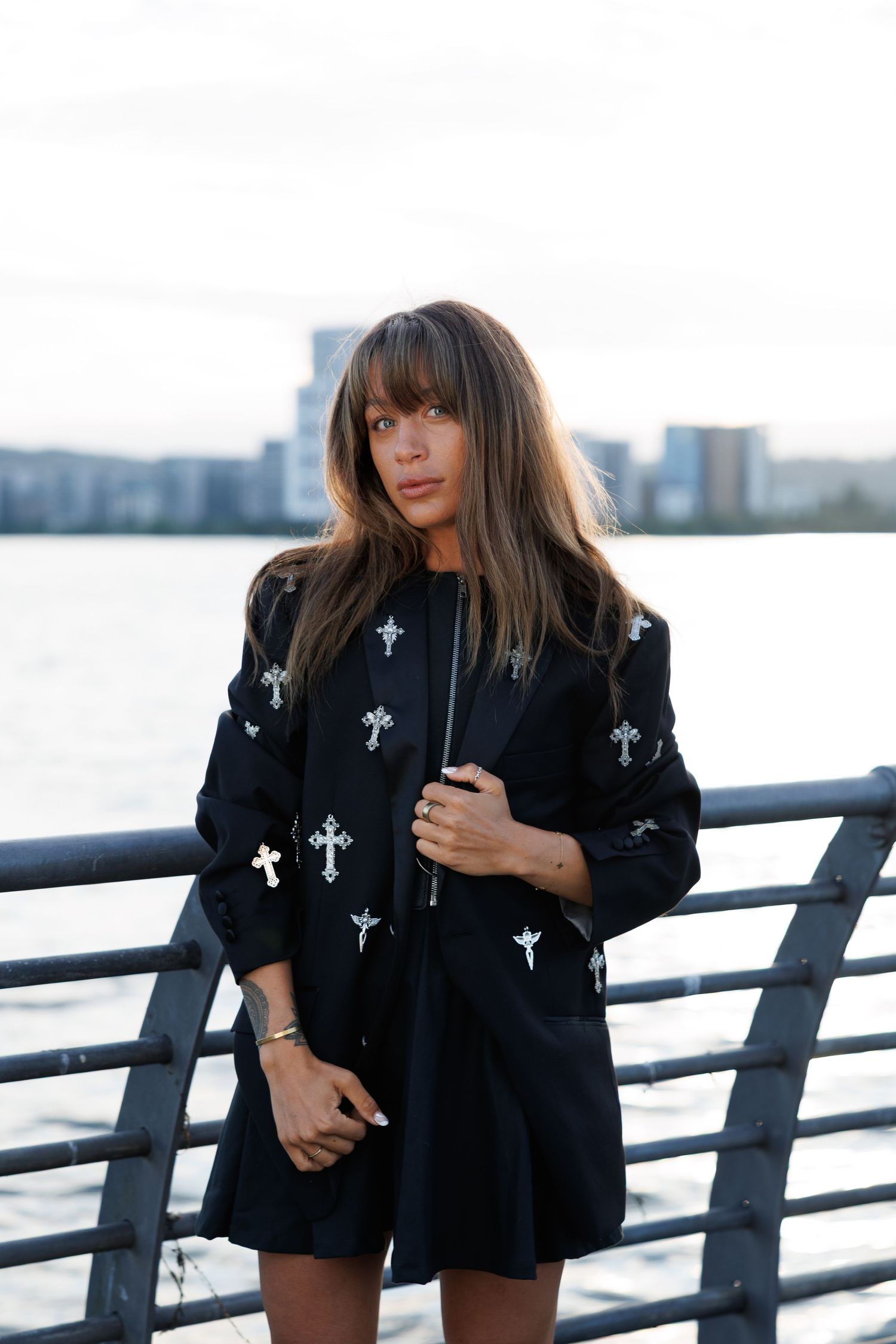 Woman in black cross-embroidered jacket posing on waterfront pier with city skyline in background.