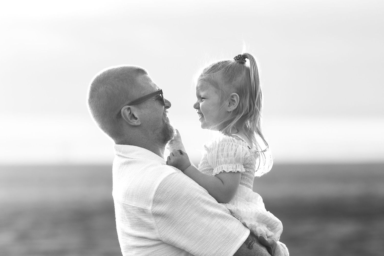 Black and white photo of a bald father holding his young daughter, sharing a tender moment outdoors.