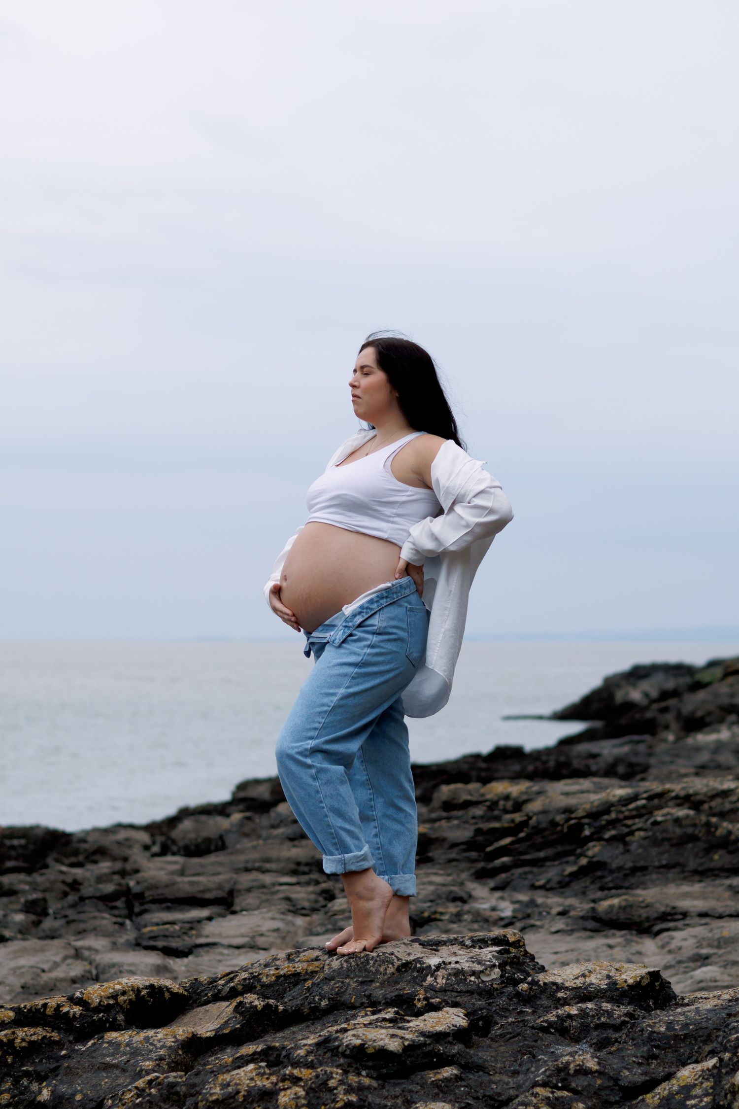 Pregnant woman in white top and jeans standing barefoot on rocky shore, cradling her baby bump by the ocean.