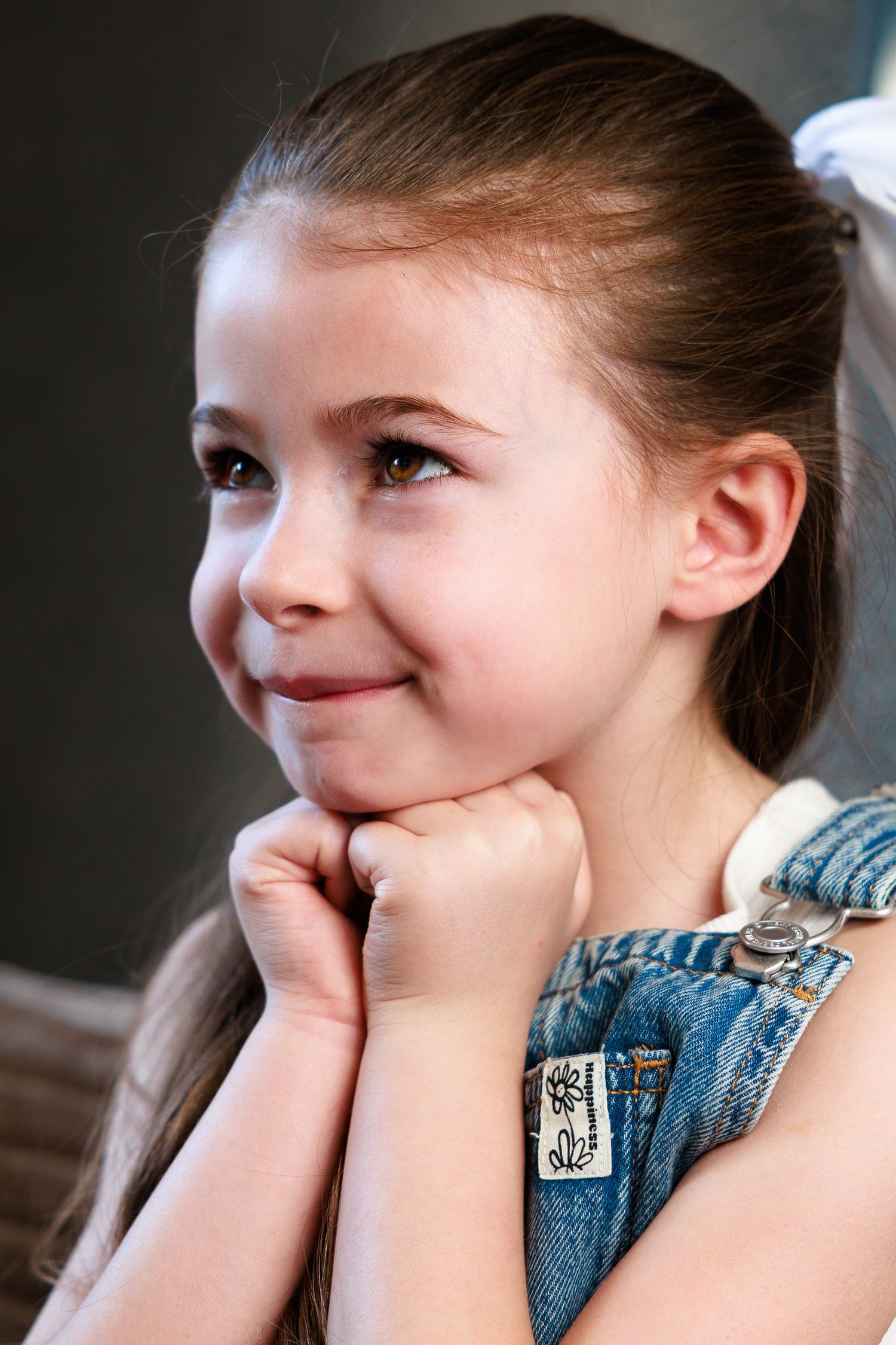 Cute young girl in denim overalls resting her chin on her hands and smiling sweetly.